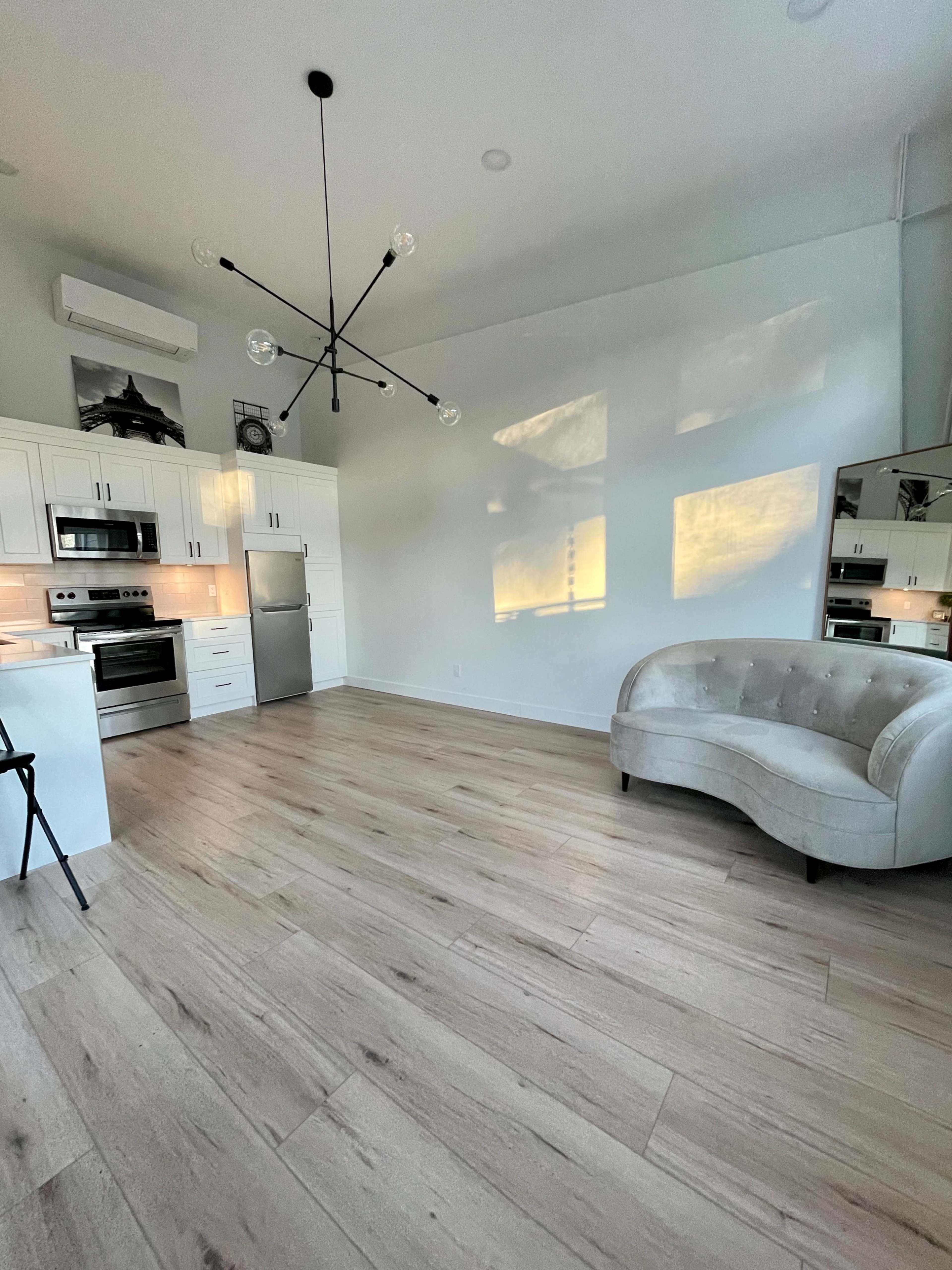 A modern kitchen with stainless steel appliances, a light-colored floor, and a tufted gray couch in a well-lit room.