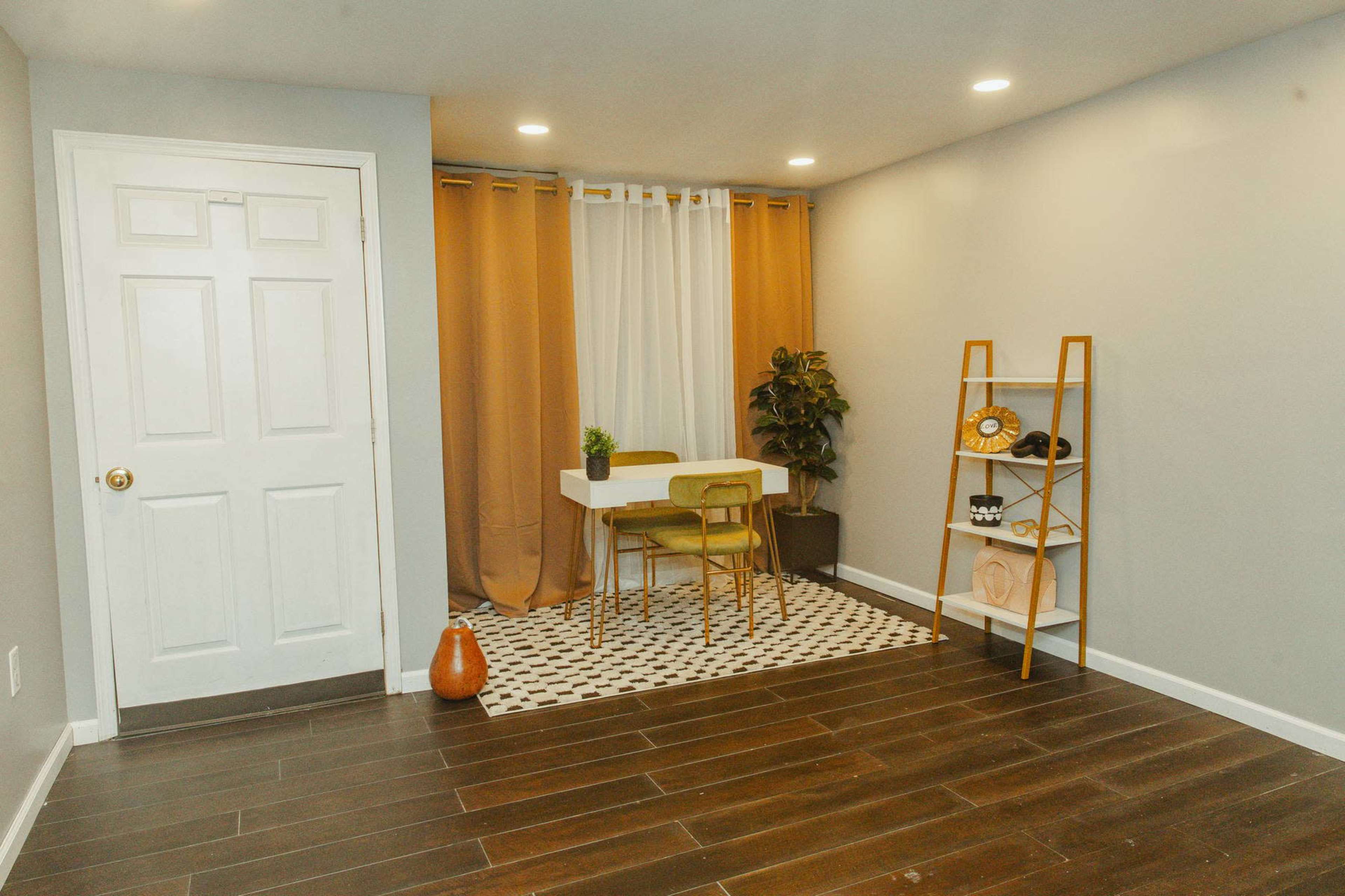 A small, well-lit room features a white table with two green chairs, a striped rug, a potted plant, and a shelving unit against a wall, with curtains and a door in the background.