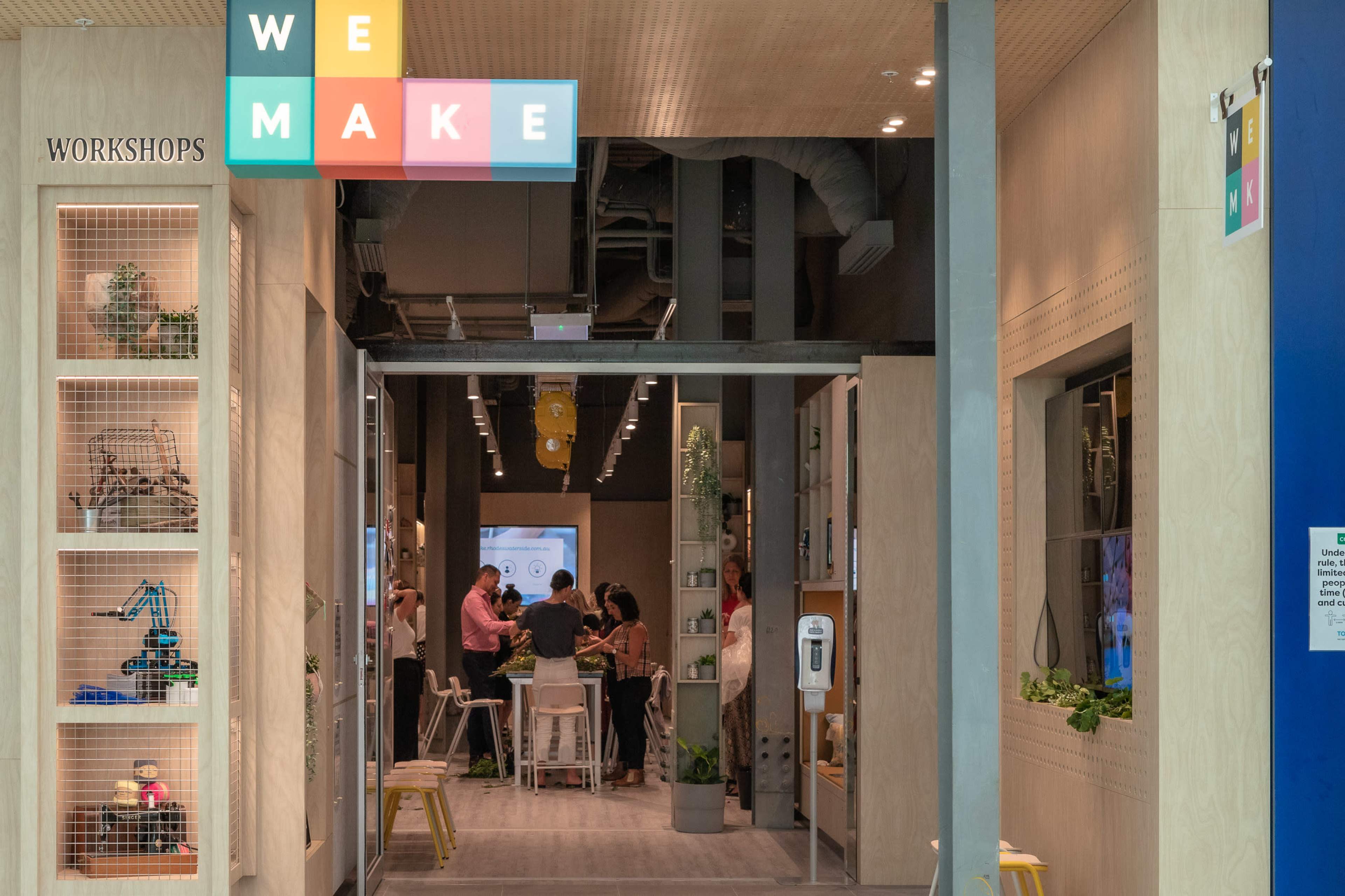 An entrance to a workshop space labeled "WE MAKE," featuring people gathered inside and various workstations along the walls.