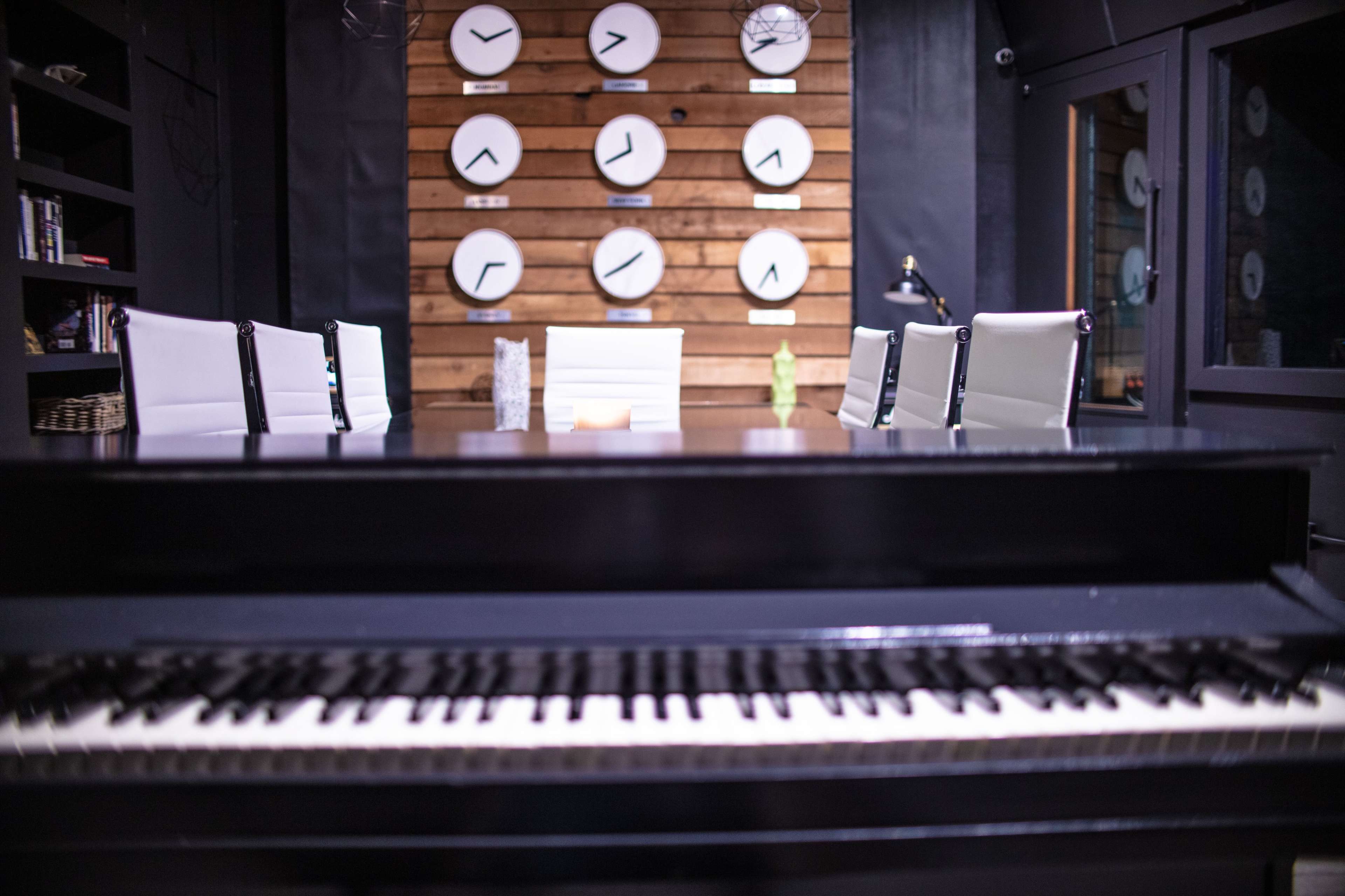 A piano sits in the foreground of a conference room featuring a long table and several clocks mounted on a wooden wall.