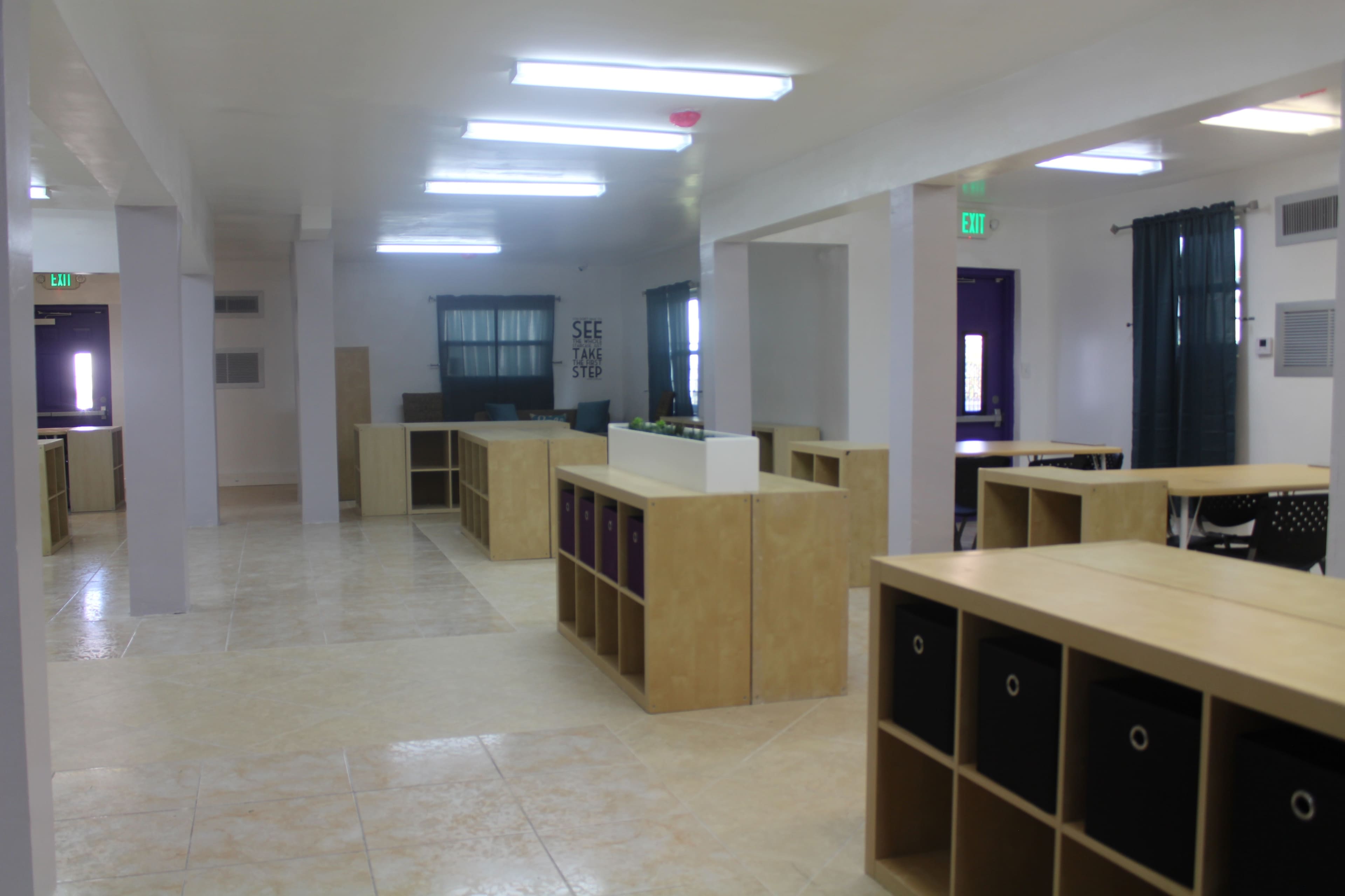The image shows a well-lit classroom or study area with several tables and shelving units arranged throughout.