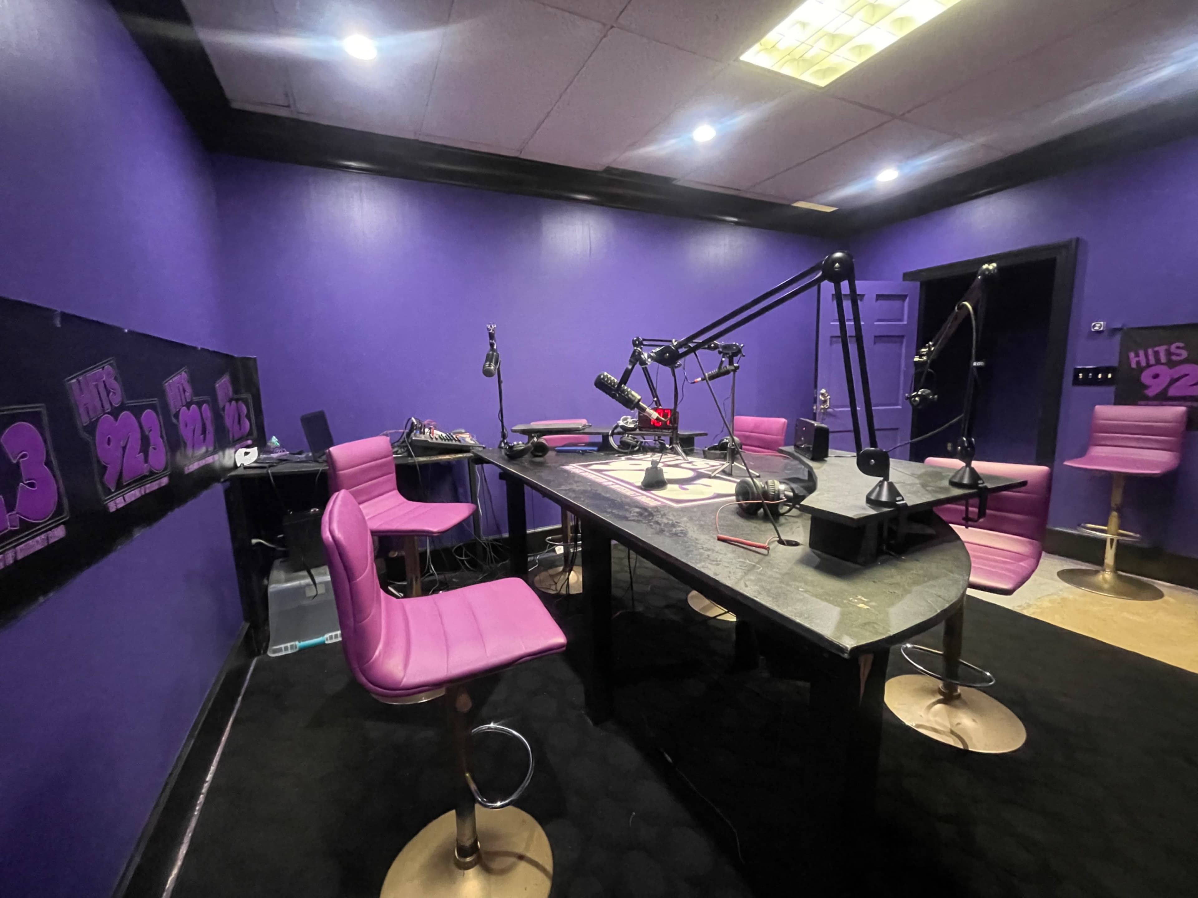 A radio studio with purple walls, a large black table equipped with microphones, and pink chairs arranged around it.