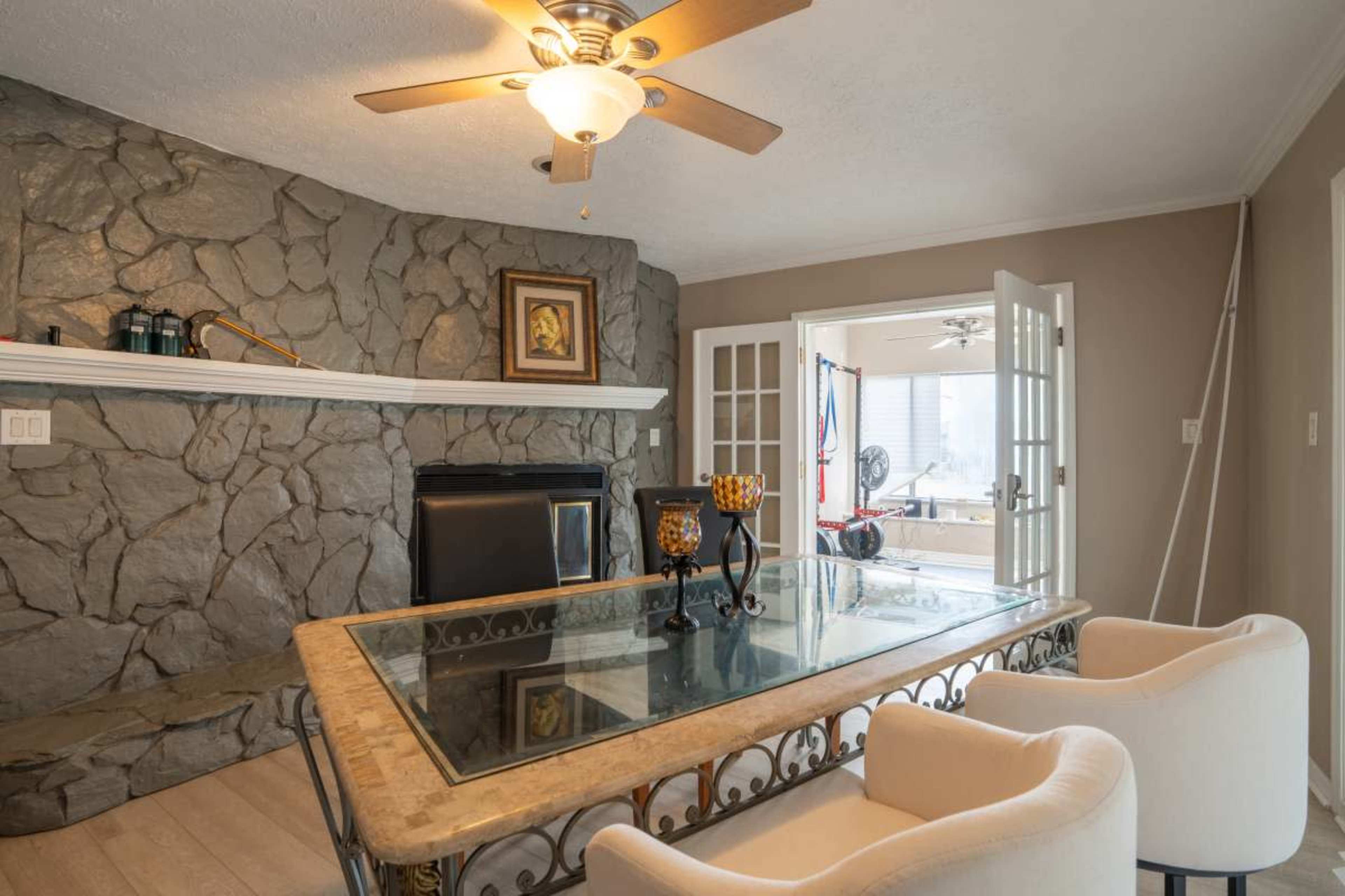 The image shows a dining area with a stone accent wall, a glass-top table, and two white chairs, leading to another room with a visible ceiling fan.