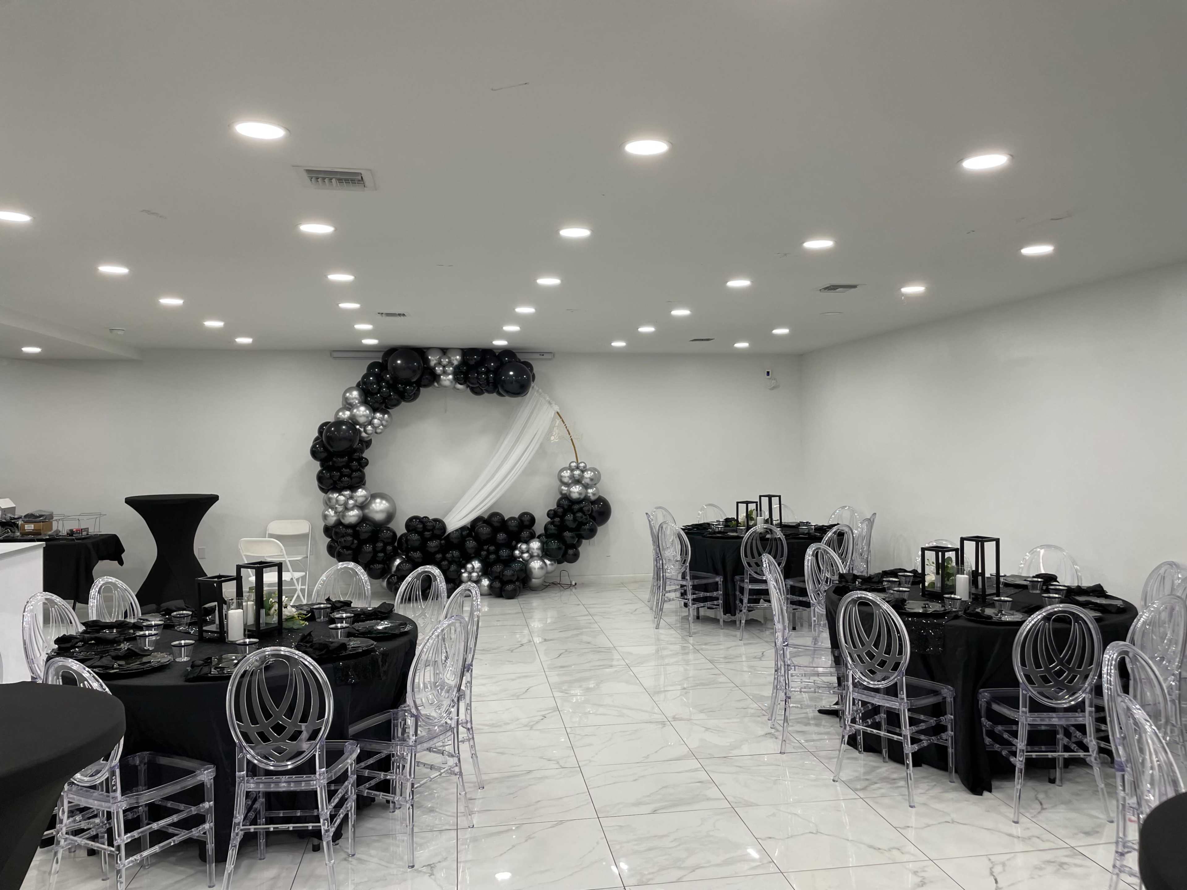 A banquet hall decorated with black and silver balloons, featuring round tables set with black tablecloths and clear acrylic chairs.