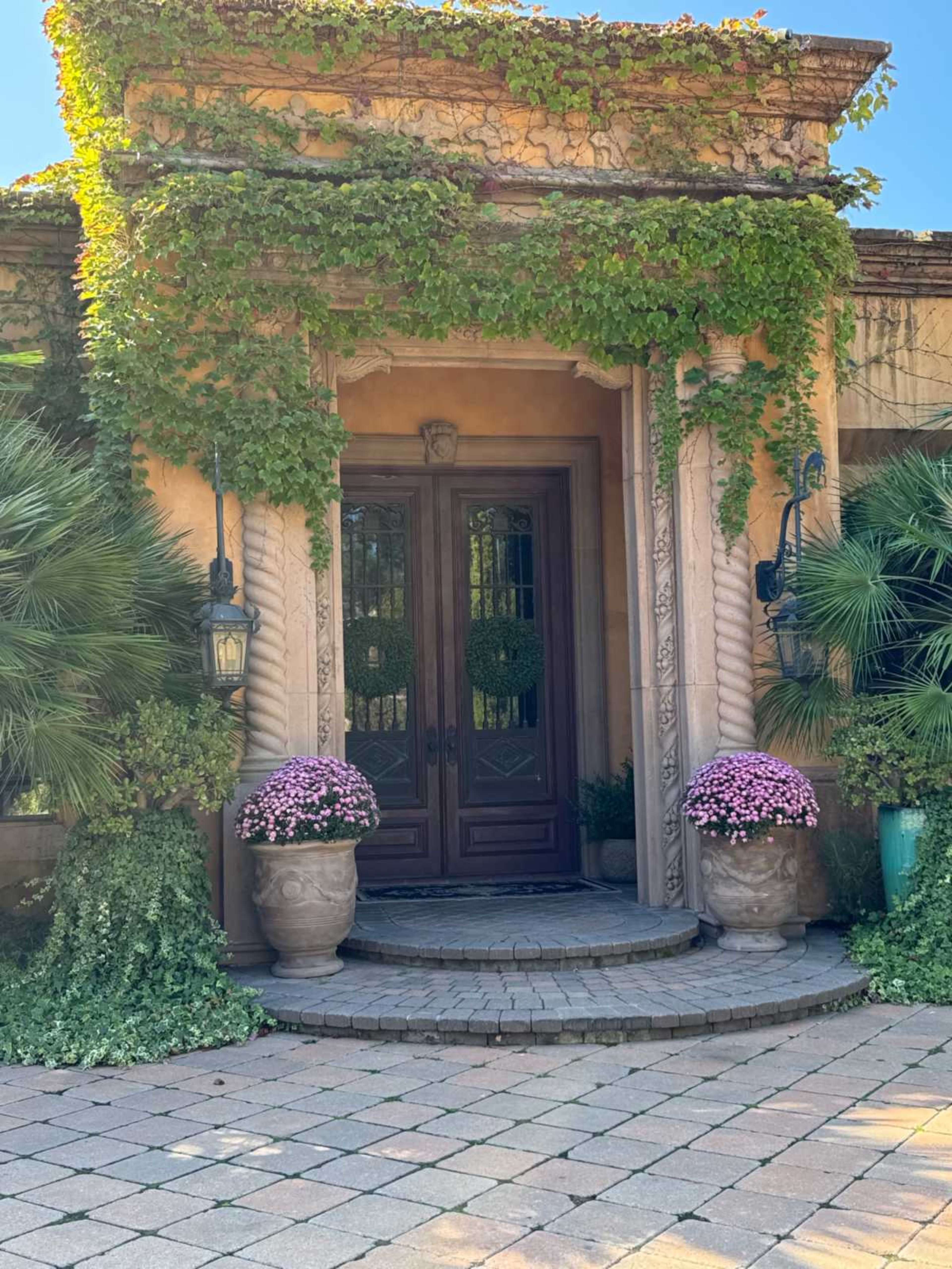 The entrance features double wooden doors framed by ornate stonework and surrounded by lush greenery and potted flowers.