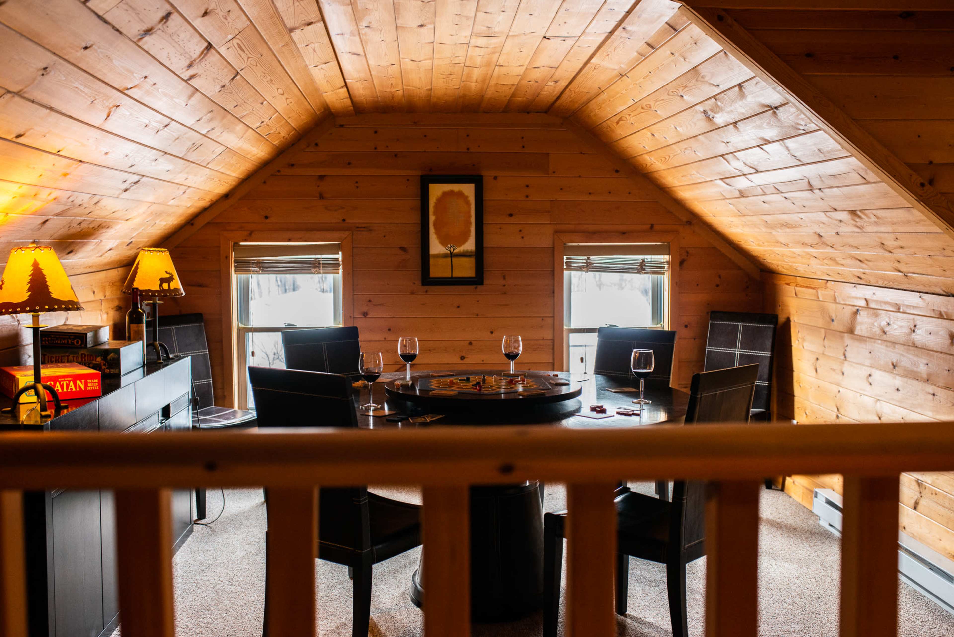 A cozy attic room features a round table surrounded by chairs, with wine glasses and board games, illuminated by warm lamps.