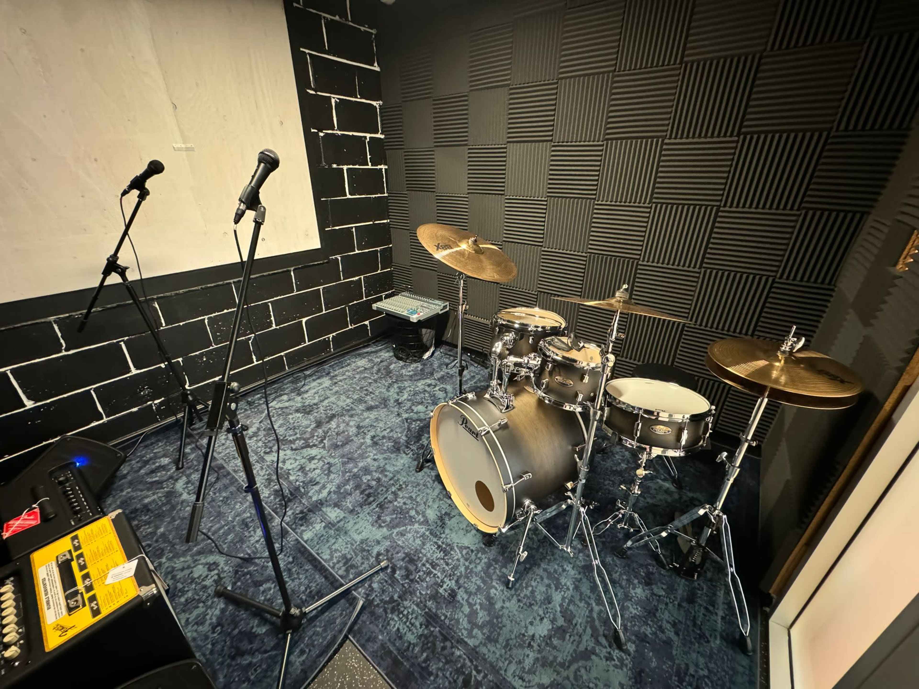 A rehearsal room featuring a drum set with cymbals, two microphones on stands, and a soundboard.
