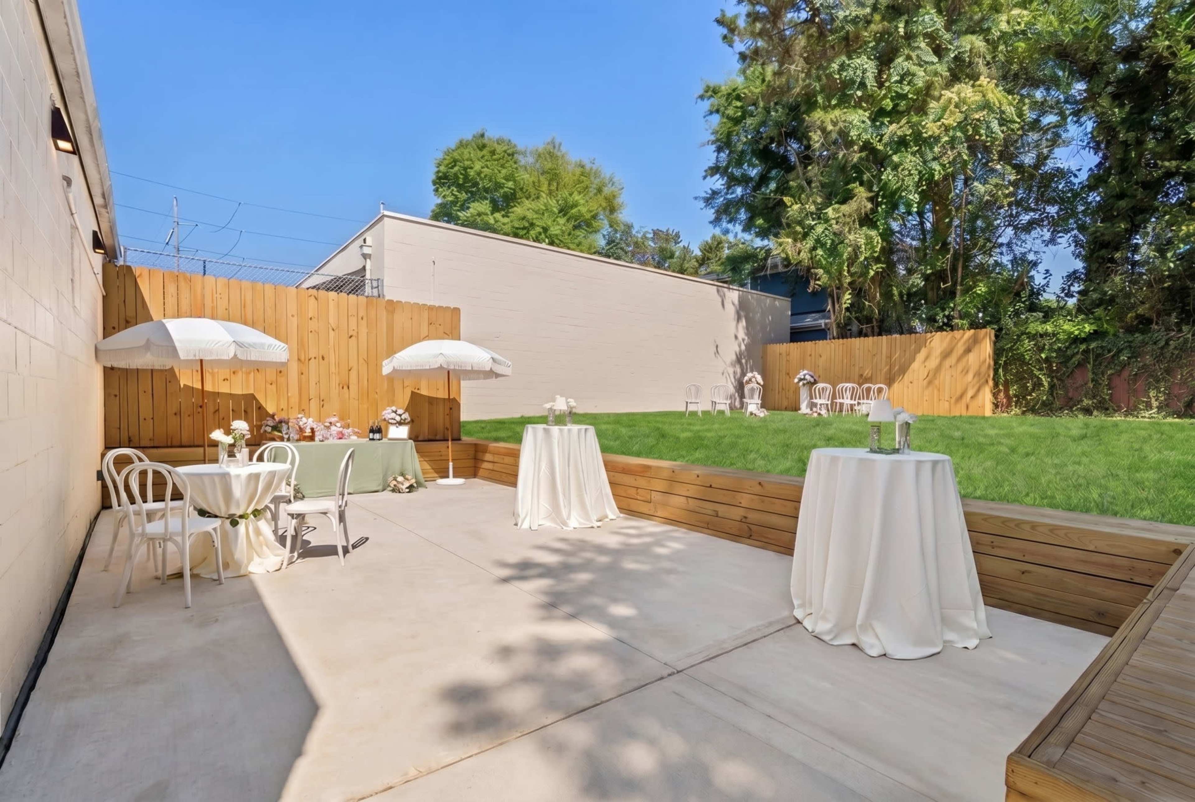 The image shows an outdoor patio area with tables covered by white tablecloths and umbrellas, surrounded by a grassy lawn and wooden fencing.