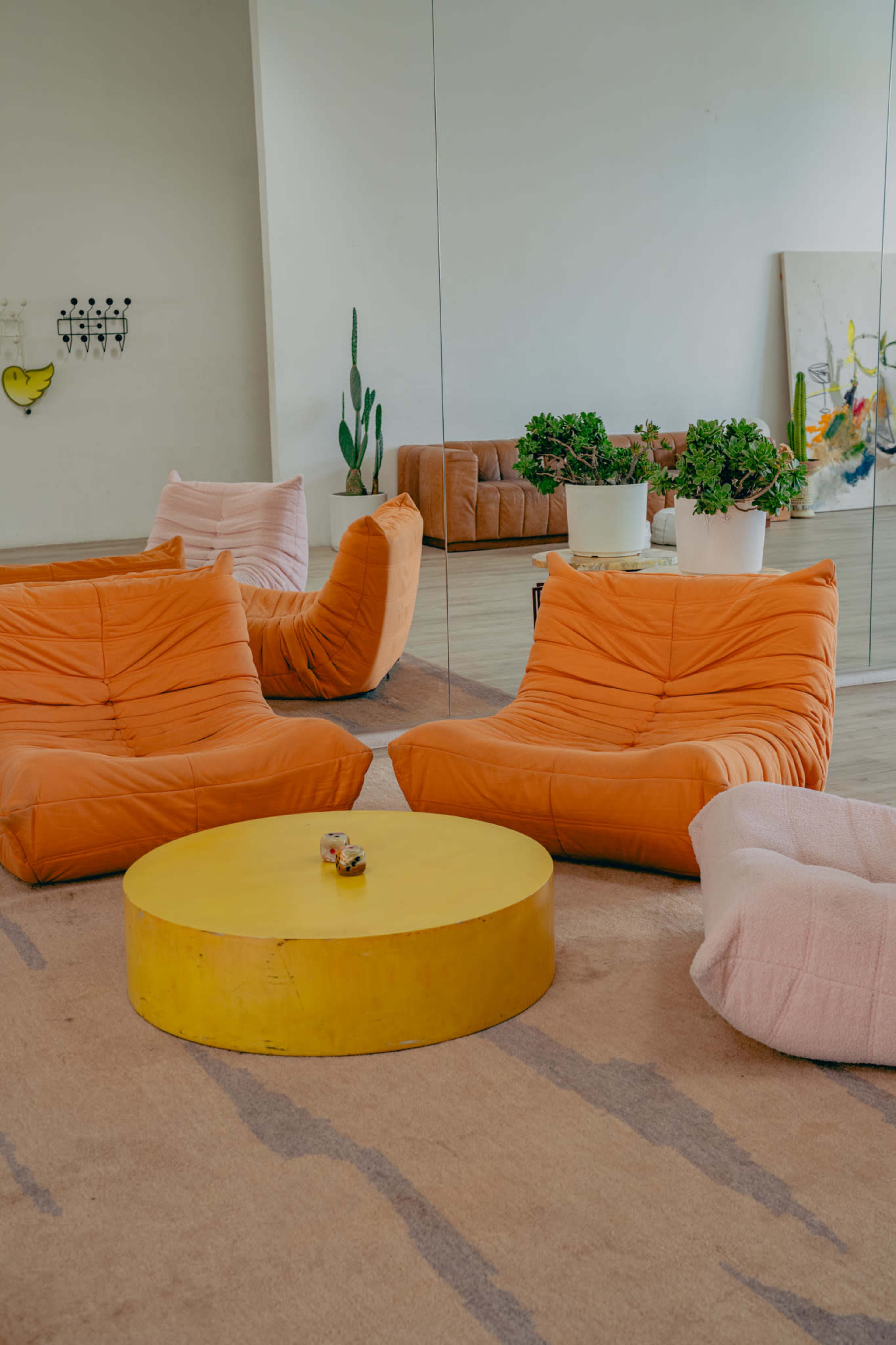 The image shows a cozy lounge area with two orange bean bag chairs, a round yellow coffee table, and potted plants, all set against a mirrored wall.