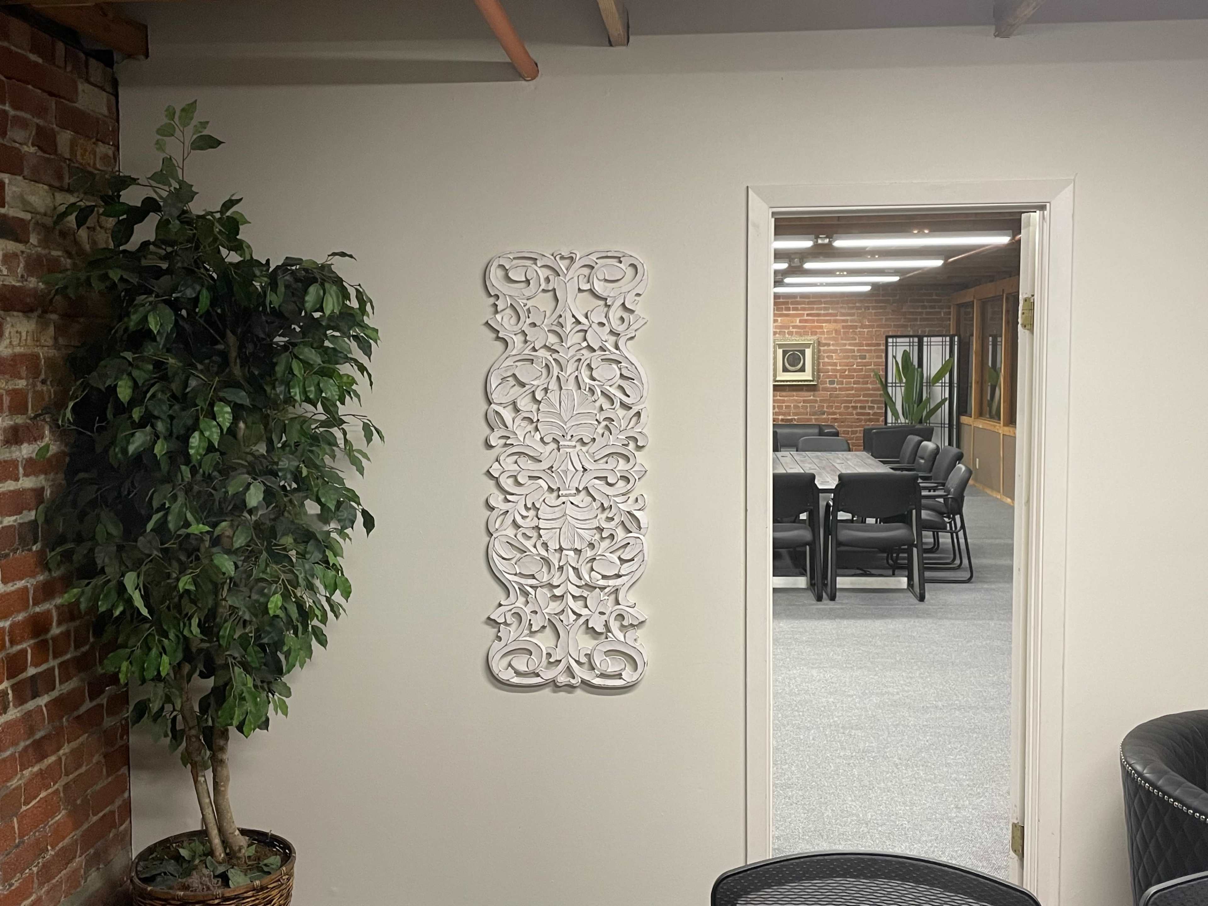 A white decorative wall panel is mounted on a light-colored wall next to a potted plant, with a doorway leading to a meeting room that features a large table and chairs.