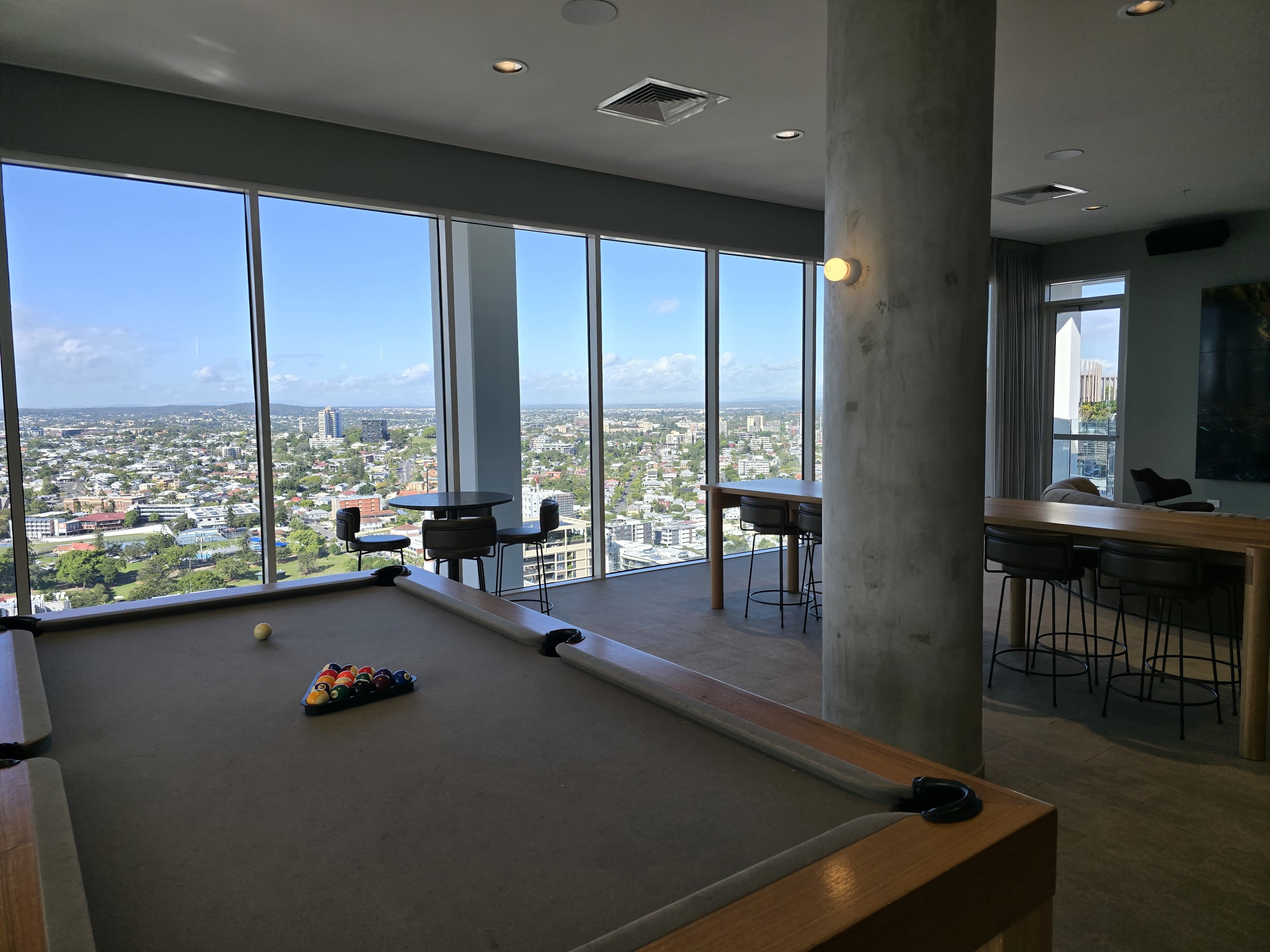 The image shows a modern interior space with a pool table in the foreground and large windows displaying a cityscape view in the background.