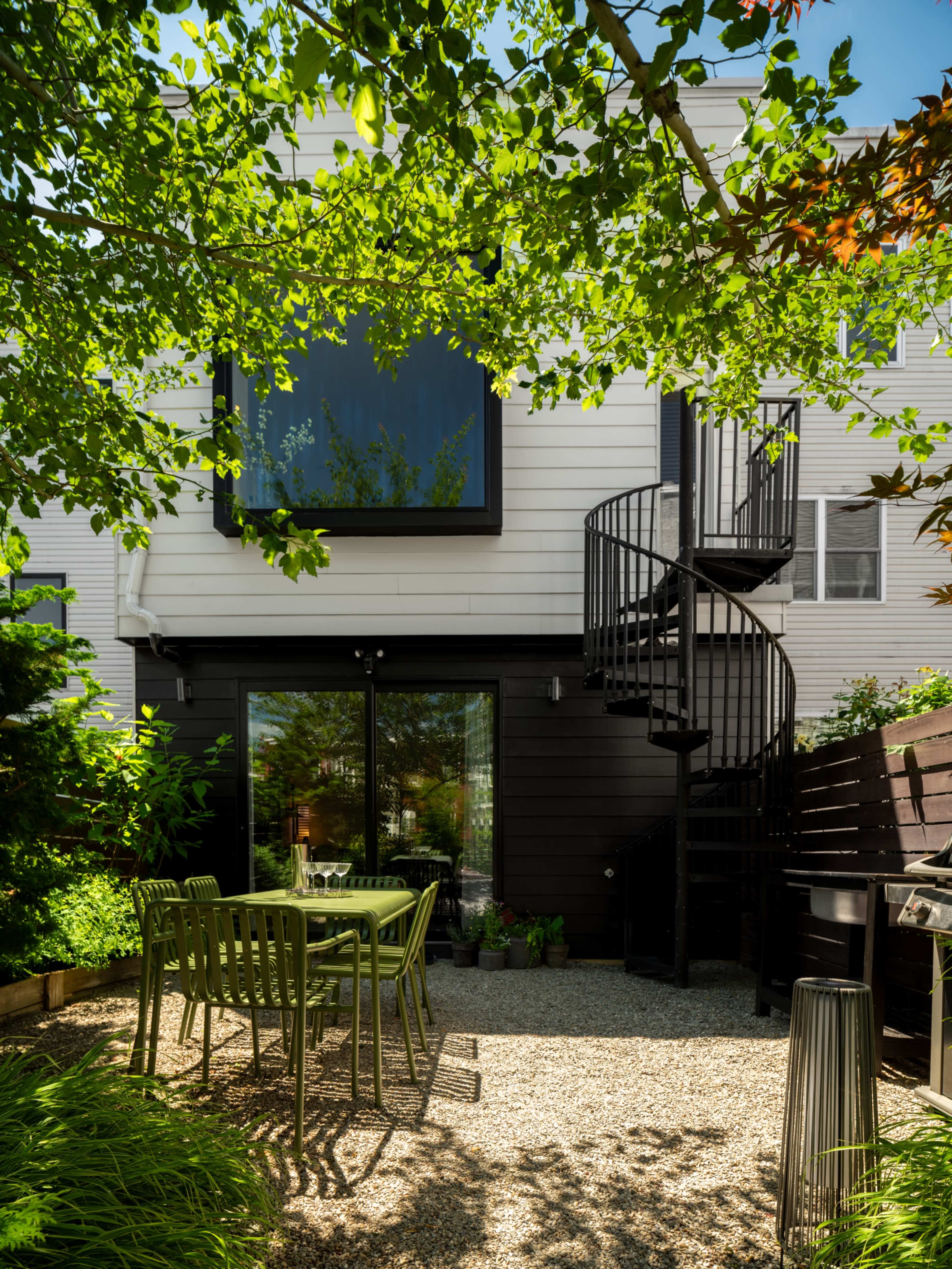A spiral staircase leads from a patio area with green chairs and a small table to the upper level of a modern home, surrounded by lush greenery.