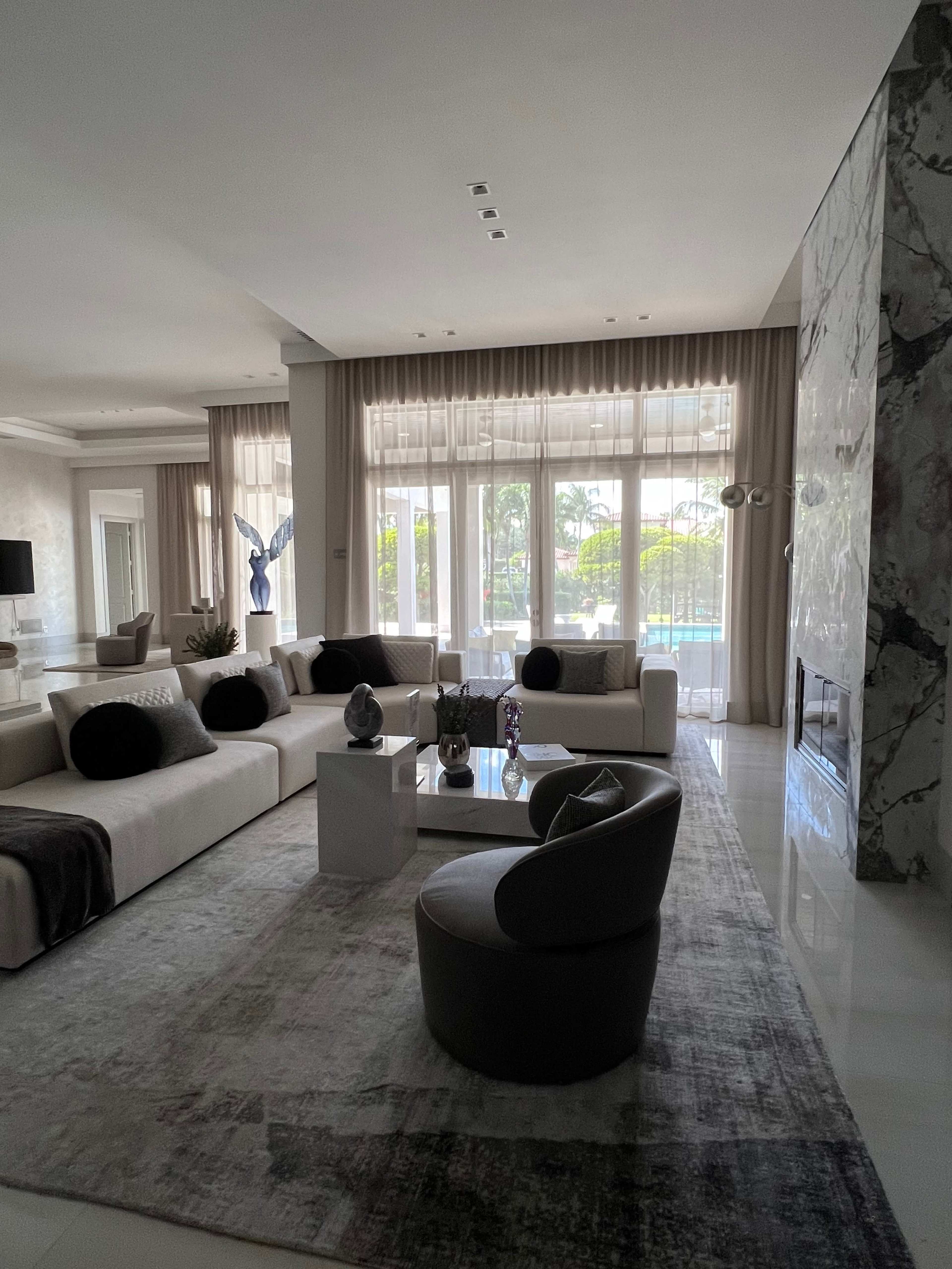 A modern living room with large windows, neutral-colored sofas, decorative cushions, a coffee table, and a rug, overlooking a green outdoor space.