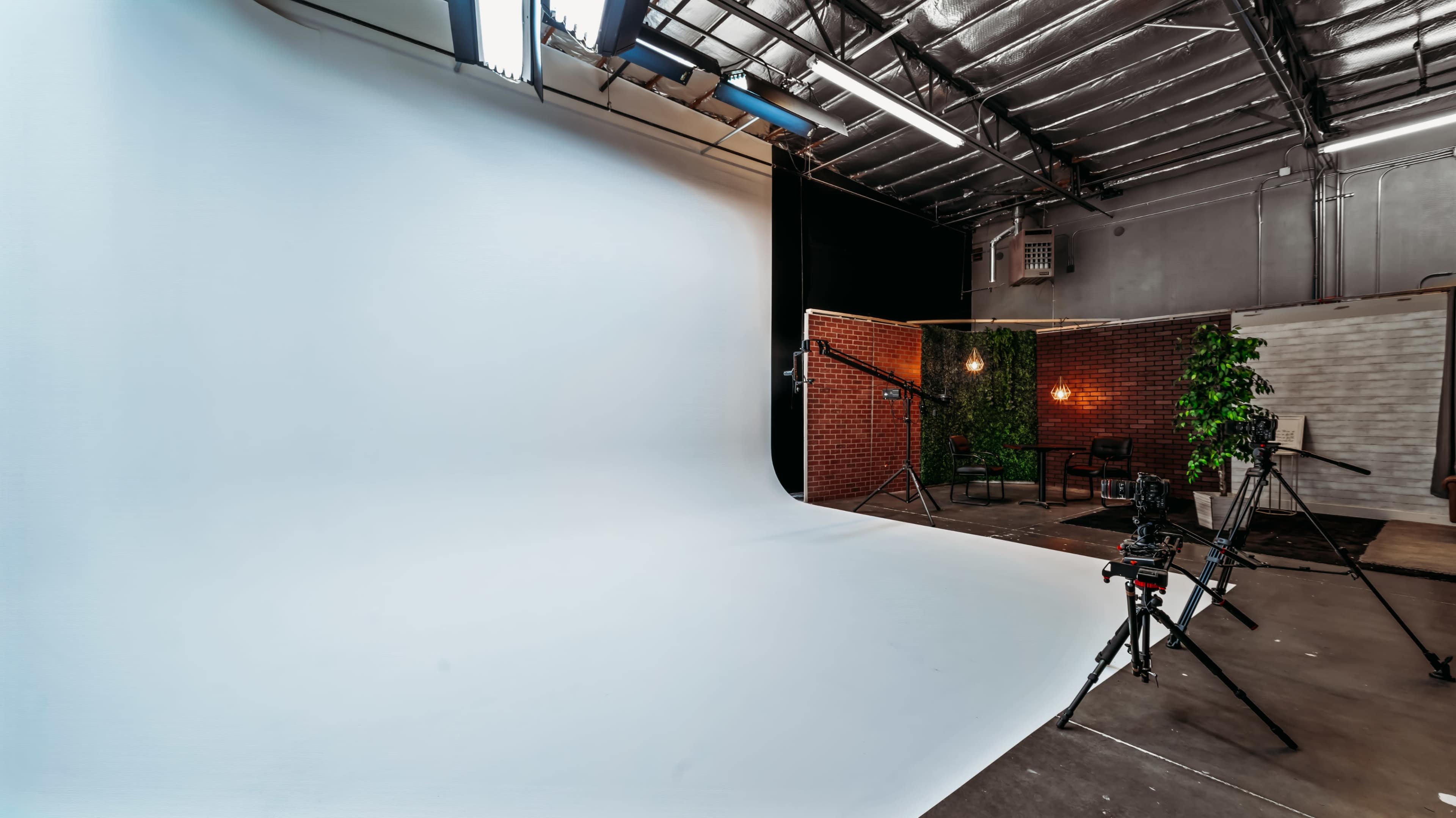 A photography studio with a white backdrop, lighting equipment, and a small seating area adorned with plants against a brick wall.