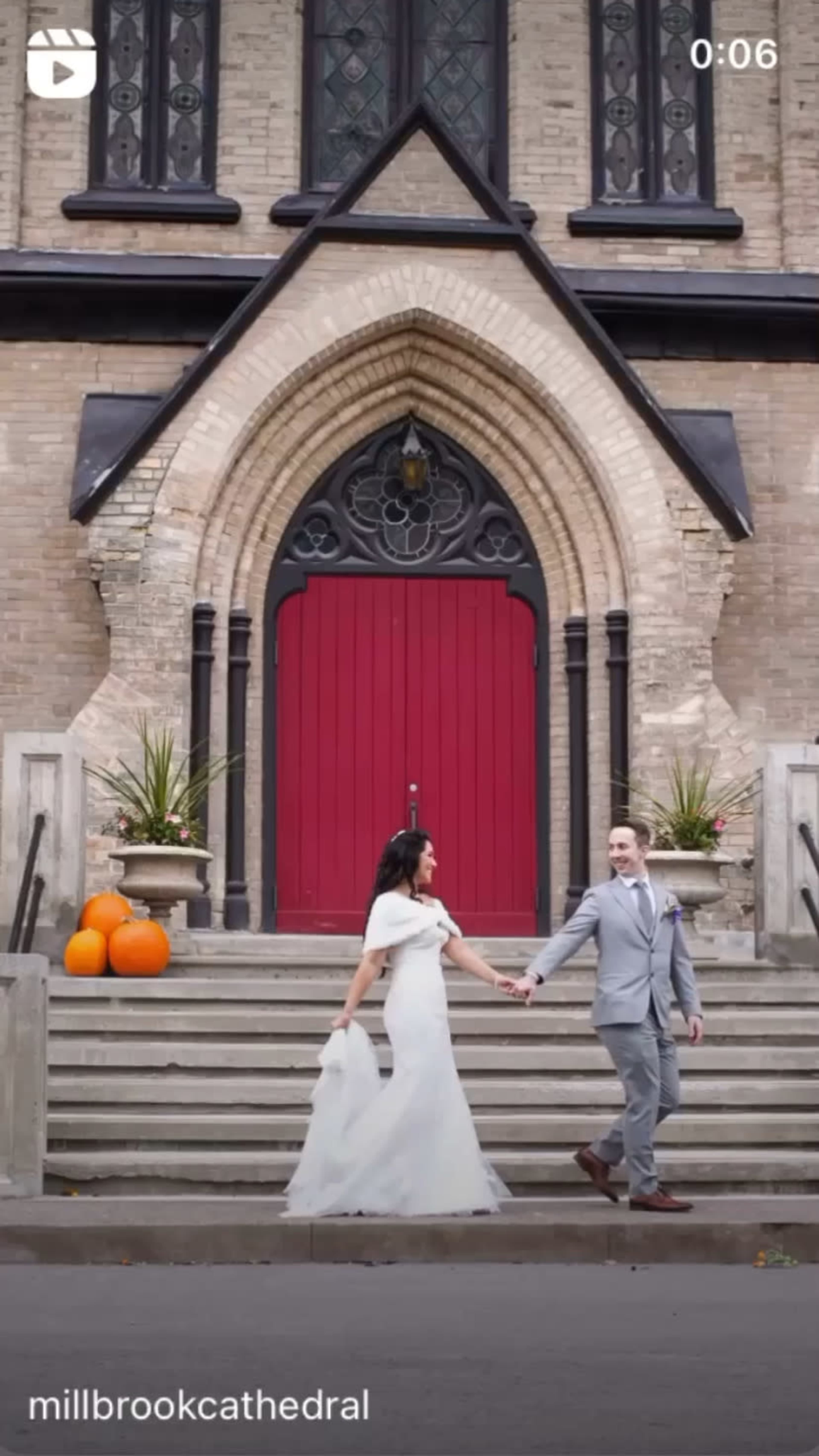 A bride and groom walk hand in hand down the steps of a cathedral, flanked by orange pumpkins.