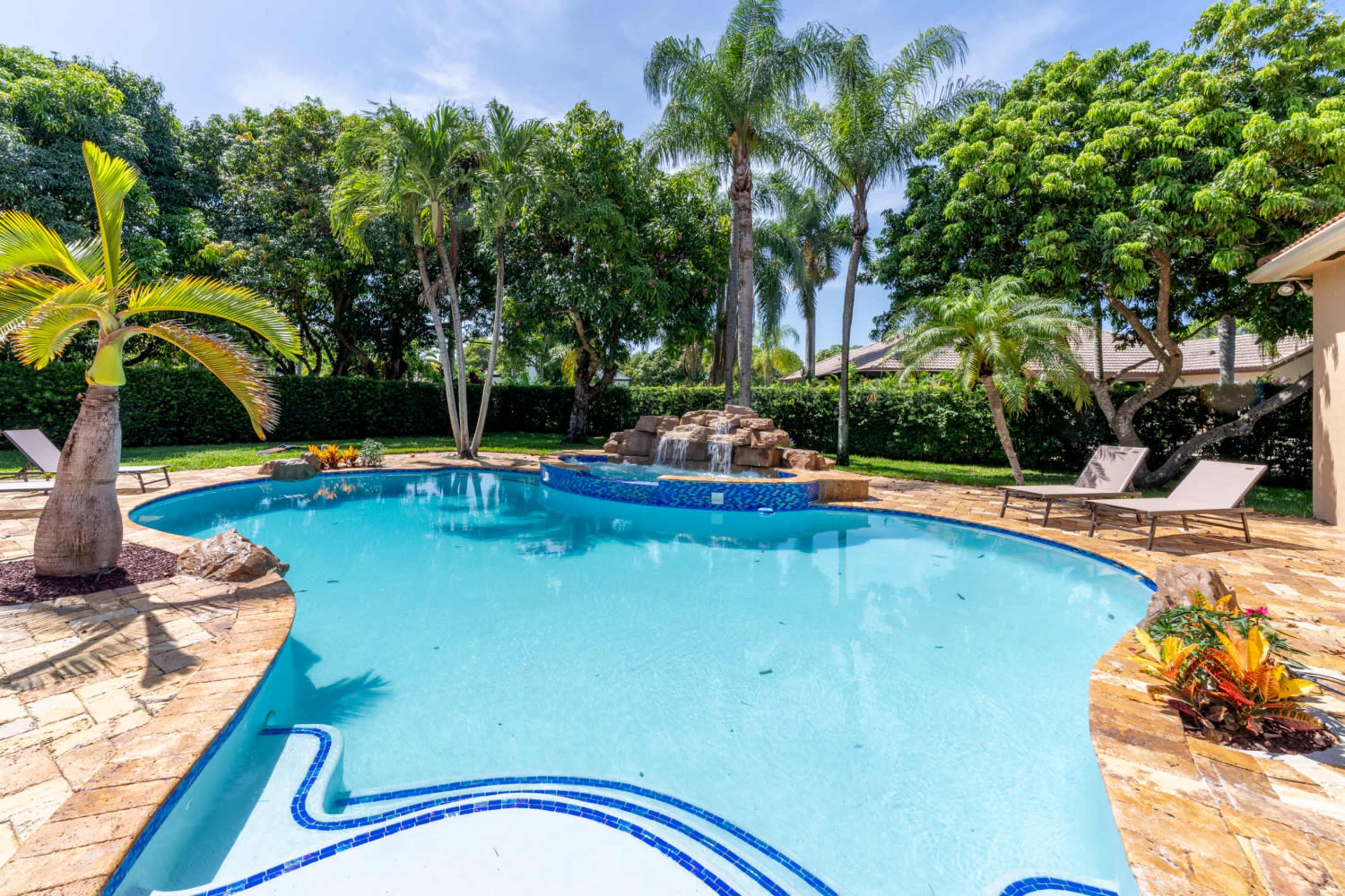 A backyard pool featuring a rock waterfall, surrounded by palm trees and lounge chairs.