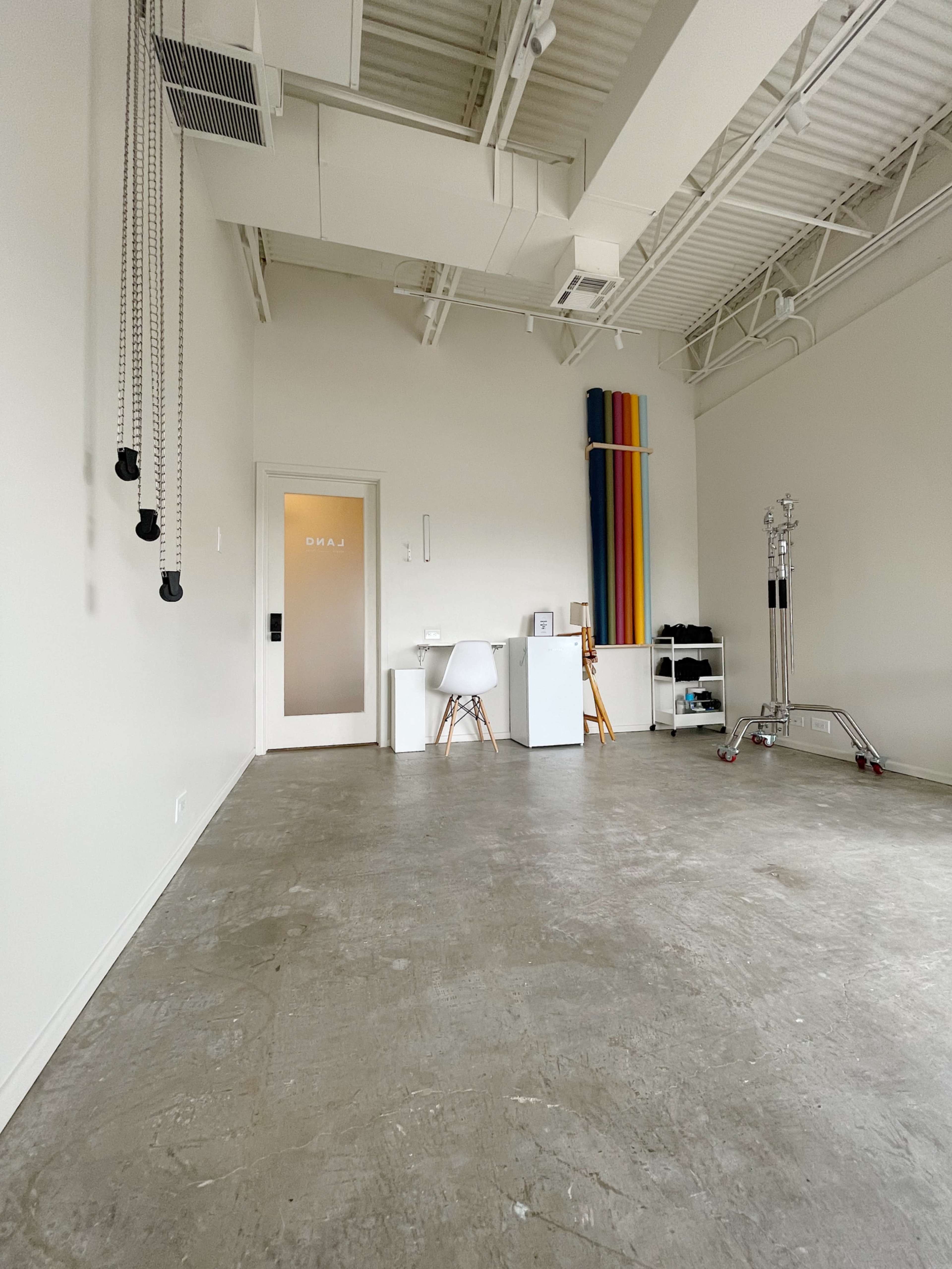 The image shows a minimalist interior space with concrete flooring, a white side table, a chair, and colorful rolls of material leaning against the wall.