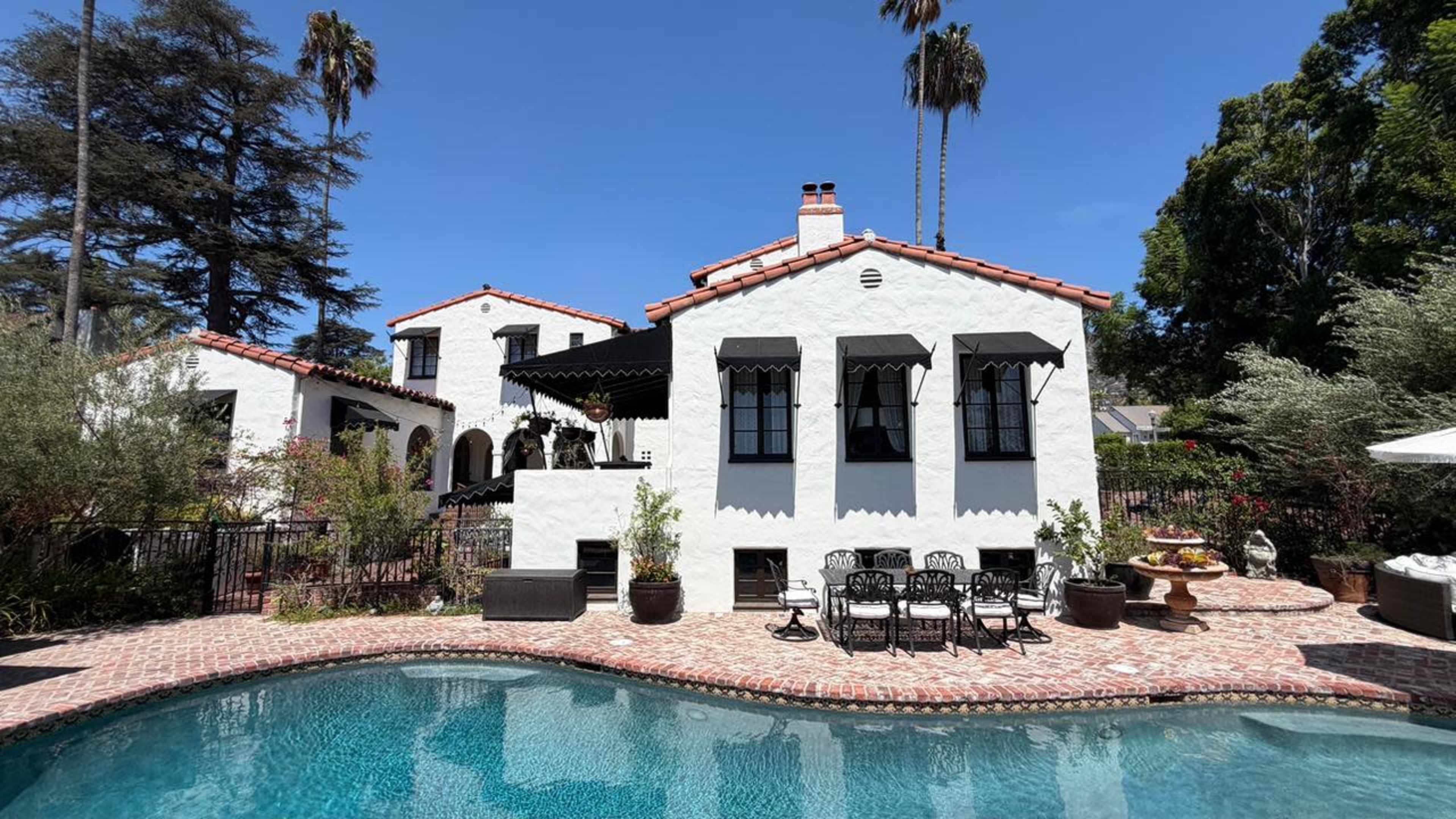 A white stucco house with black window awnings and a red tile roof is situated beside a swimming pool surrounded by lush greenery and decorative plants.