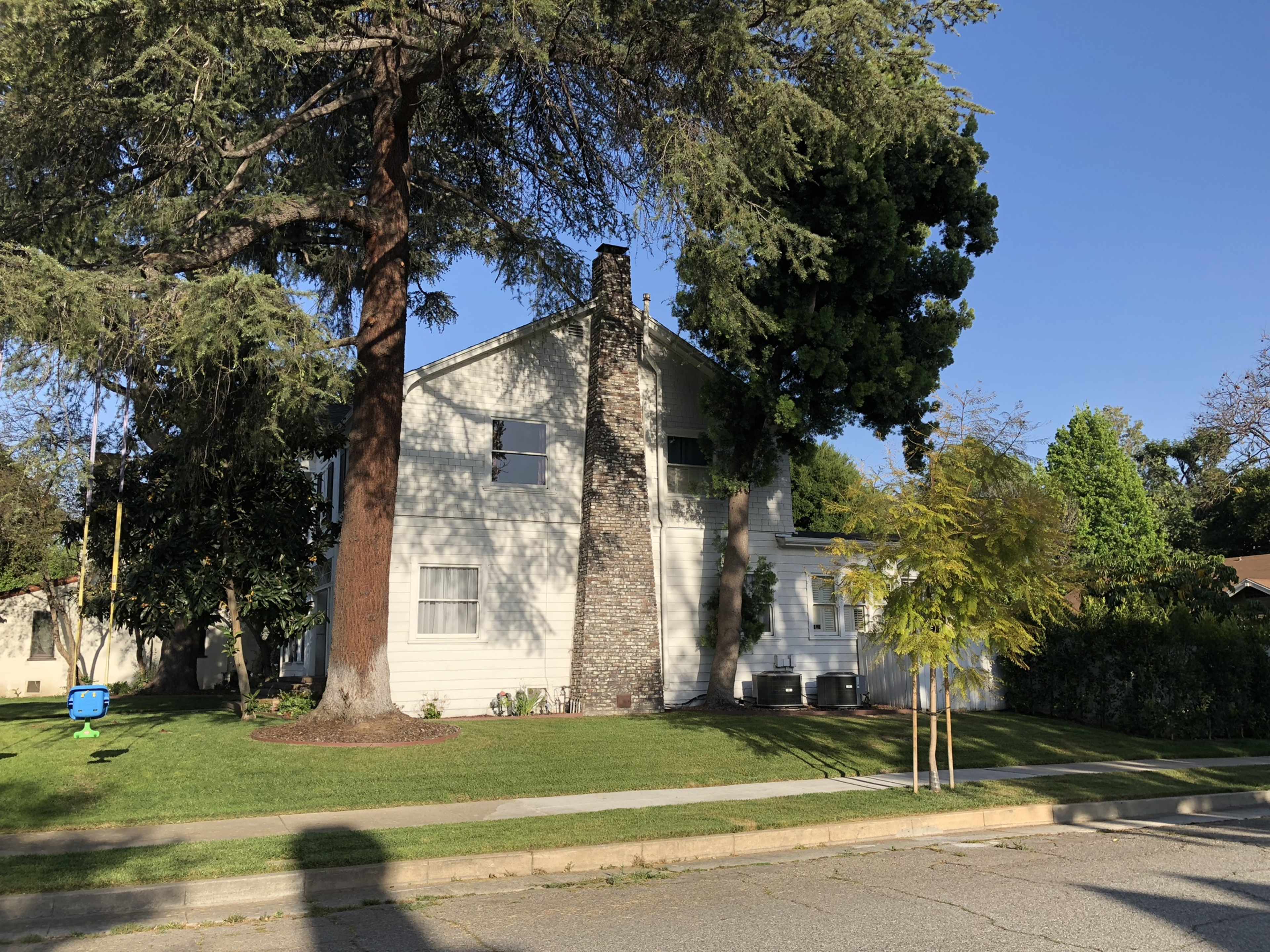 A two-story house with a stone chimney is surrounded by trees and grass on a residential street.