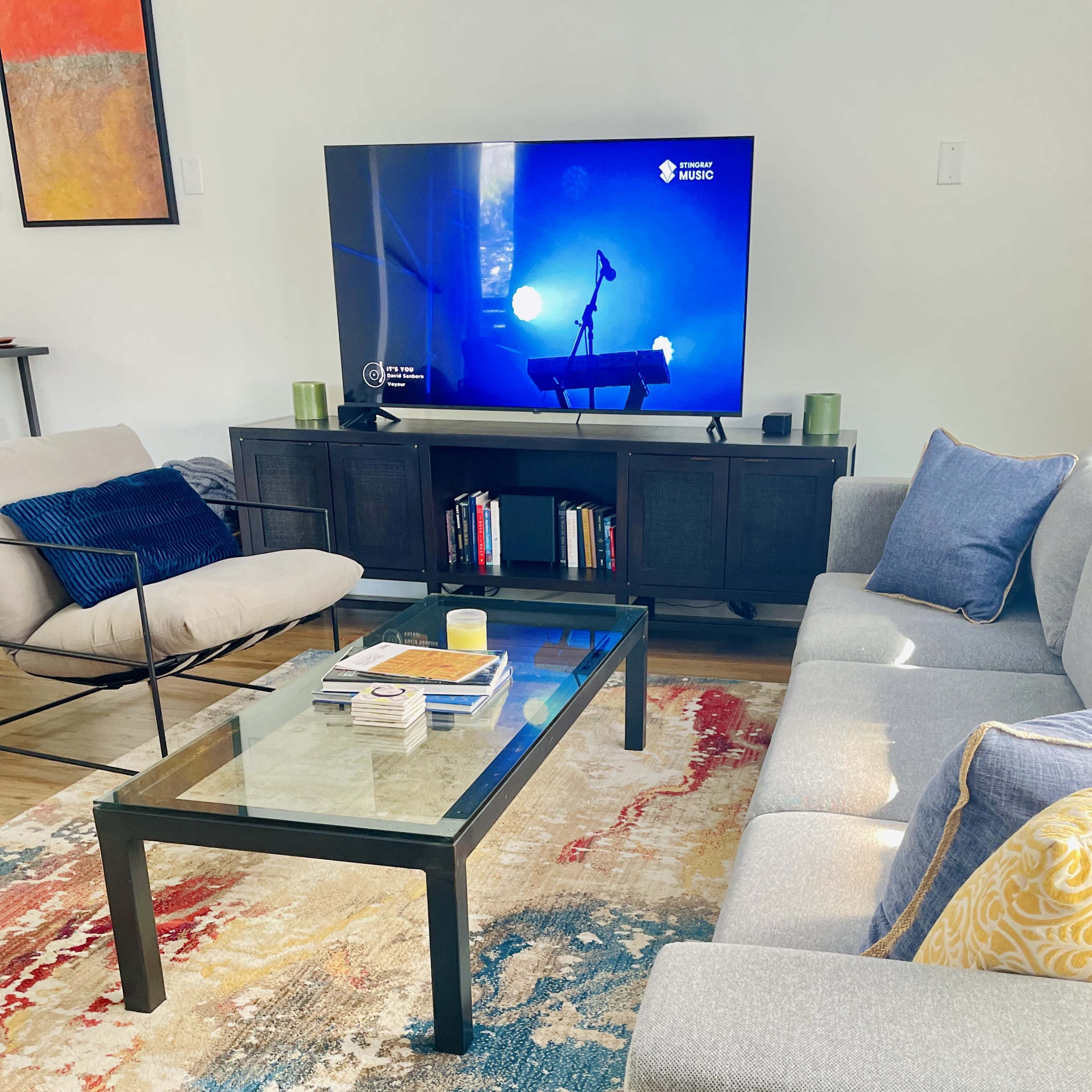 The image shows a living room with a gray sofa, a glass coffee table, a black media console with books, and a television displaying a blue-lit scene.