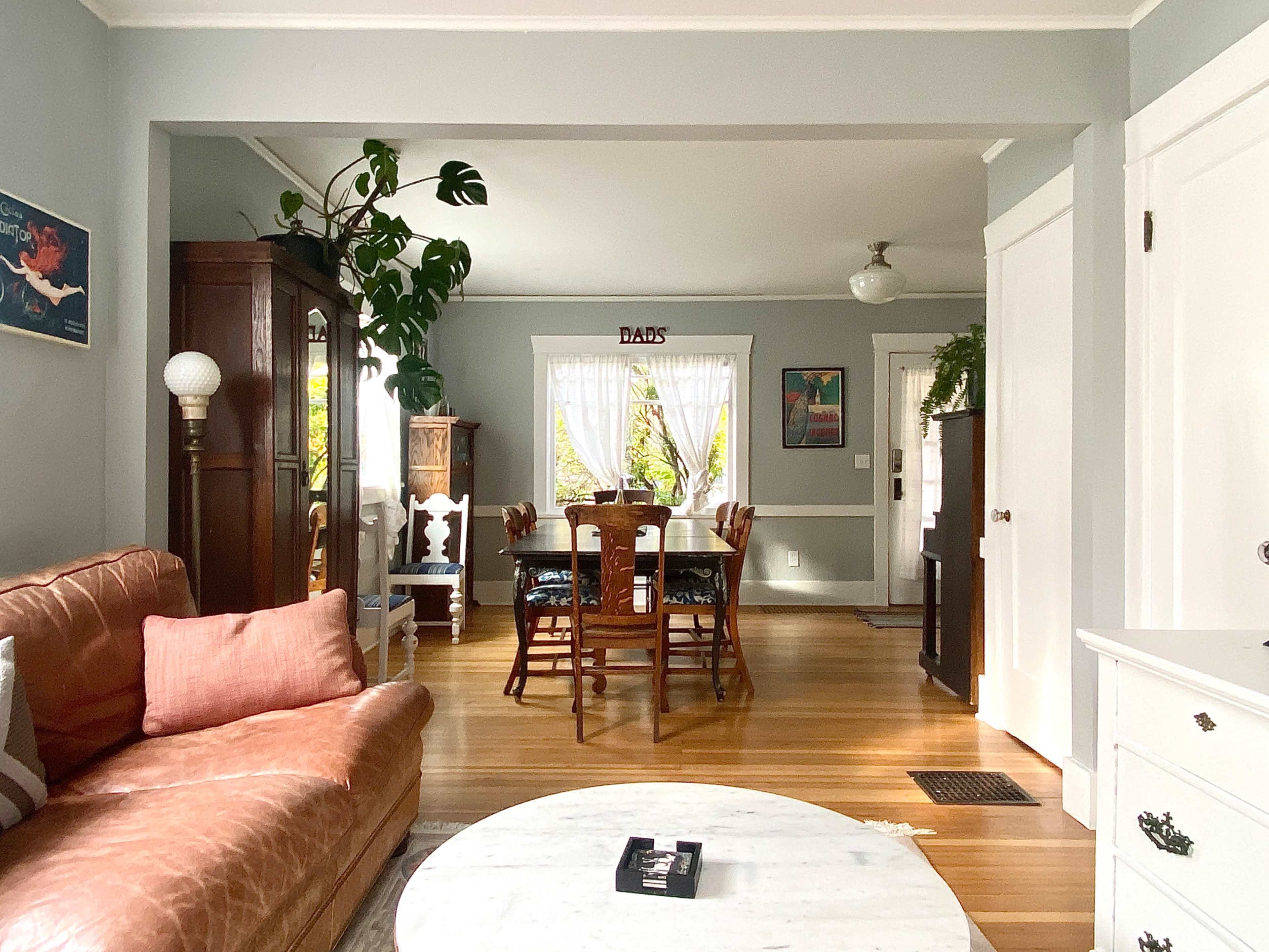 A cozy living room leads to a dining area with wooden furniture and large windows letting in natural light.