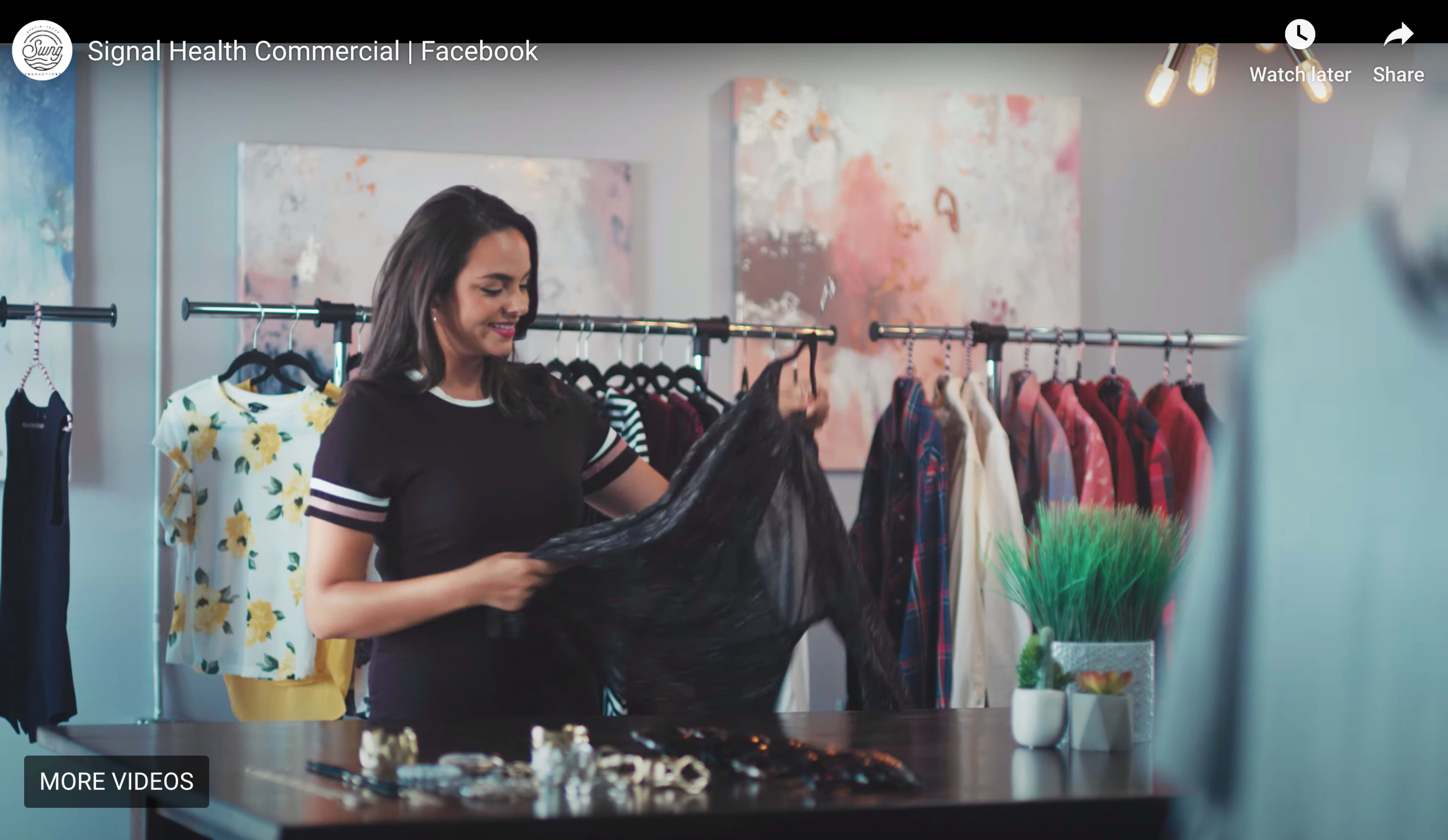 A woman is inspecting a black garment while standing at a table in a clothing store filled with hanging clothes.