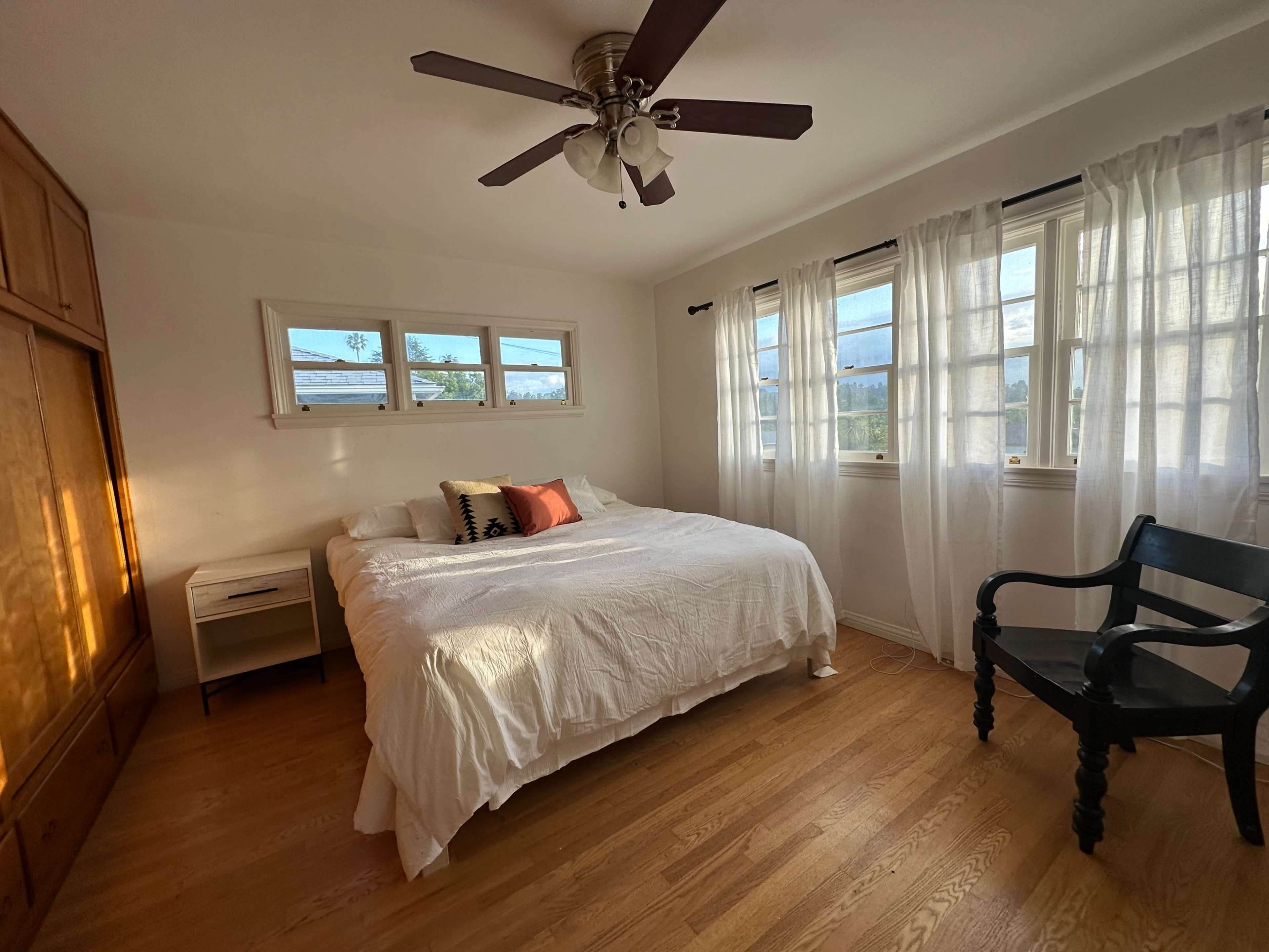 The image shows a bright bedroom with a large bed, a wooden chair, and a wall of windows covered by sheer curtains.