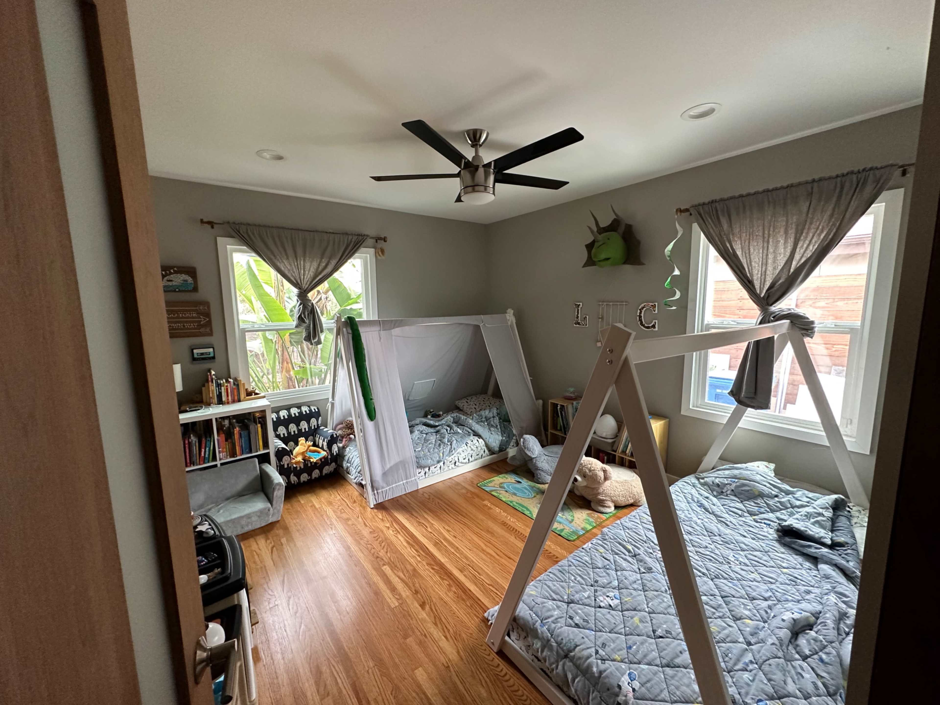 A children's bedroom with two beds, a small bookshelf, and a ceiling fan, all arranged around a window with white curtains.