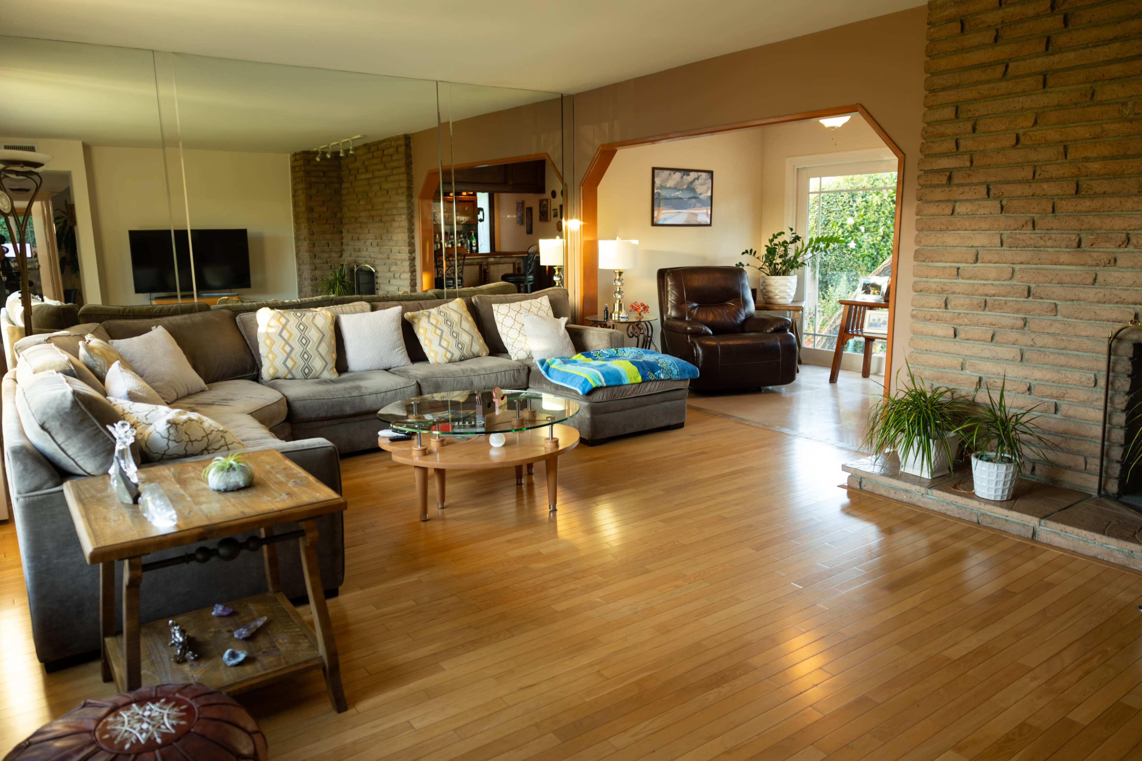 A spacious living room featuring a large sectional sofa, a wooden coffee table, and a seating area with a recliner, surrounded by large windows and plants.