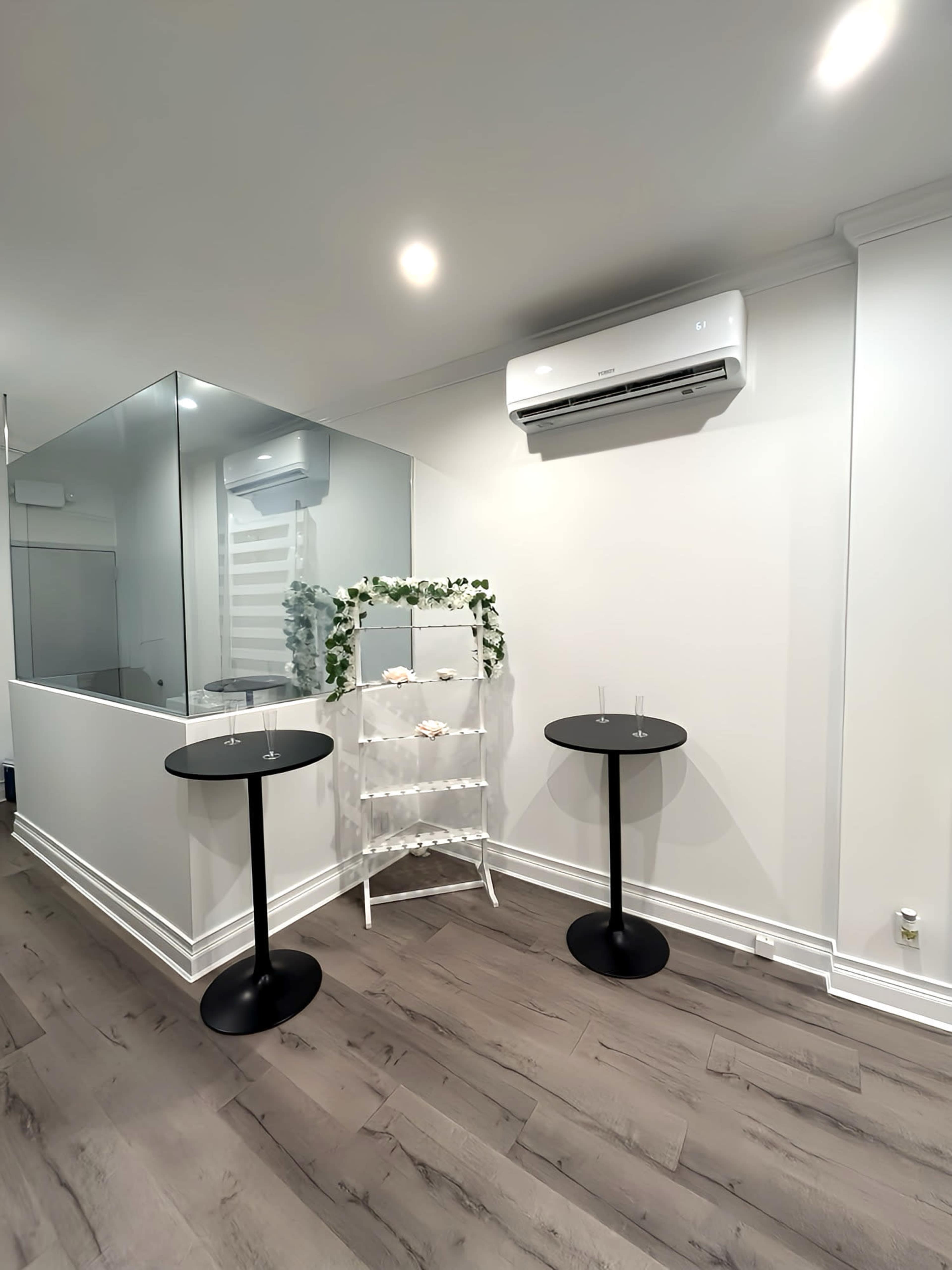 The image shows a modern, minimalistic interior with two black tables, a glass partition, and a decorative shelf adorned with greenery.