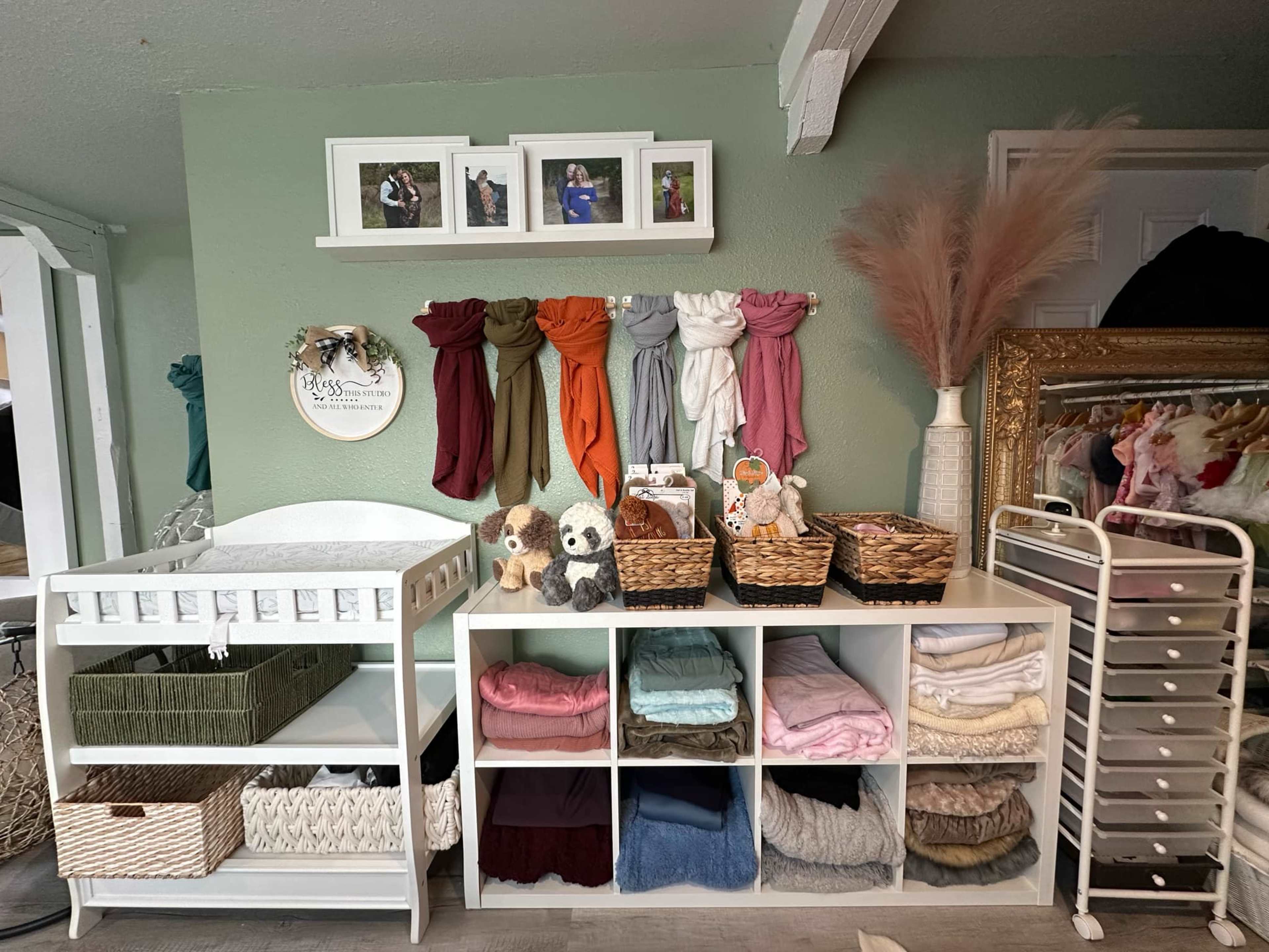 The image shows a neatly organized nursery setup with a changing table, shelves of folded blankets, and decorative items including framed photos and storage baskets.