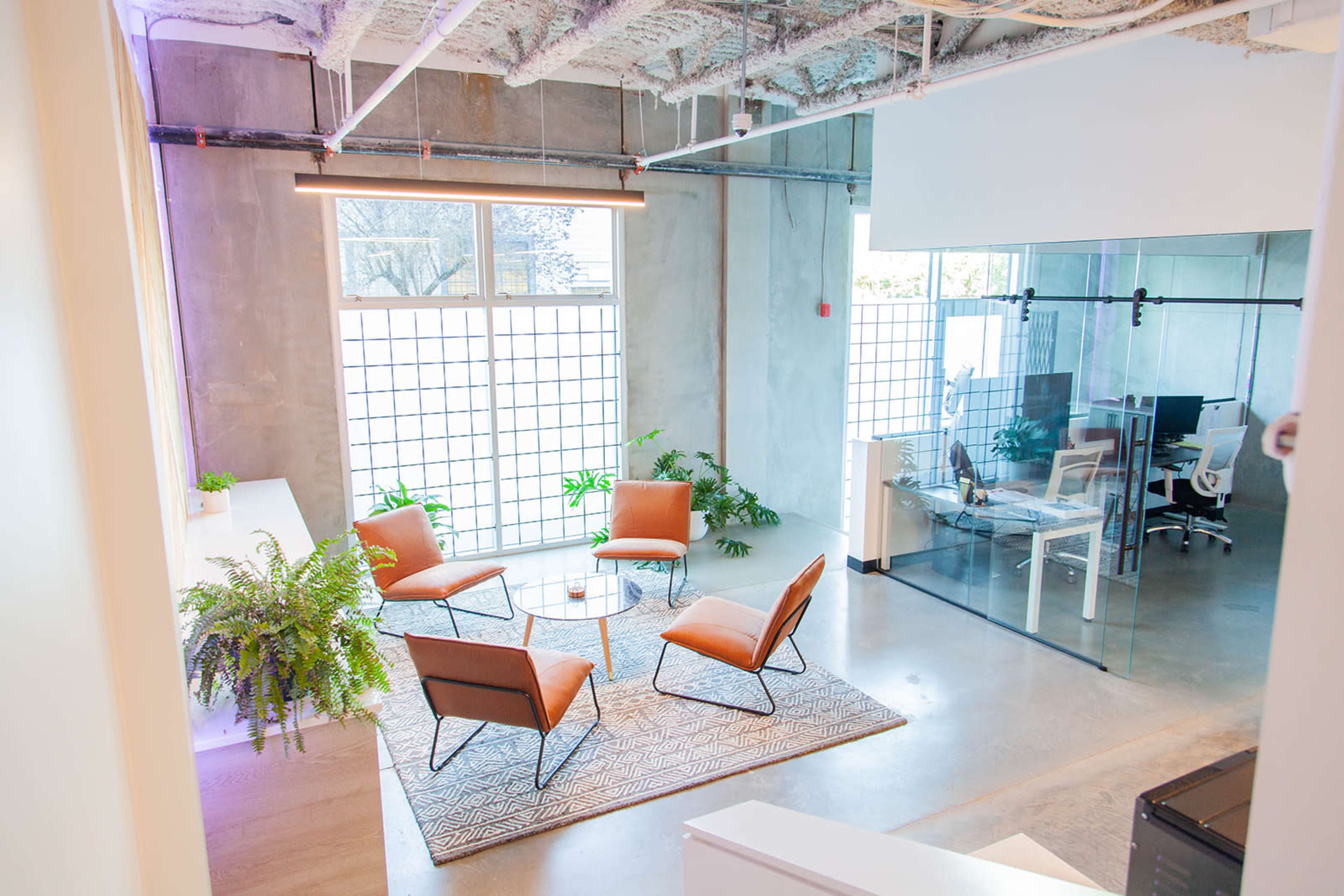 A modern office space featuring a seating area with four orange chairs, a glass partitioned workspace, and natural light coming through large windows.