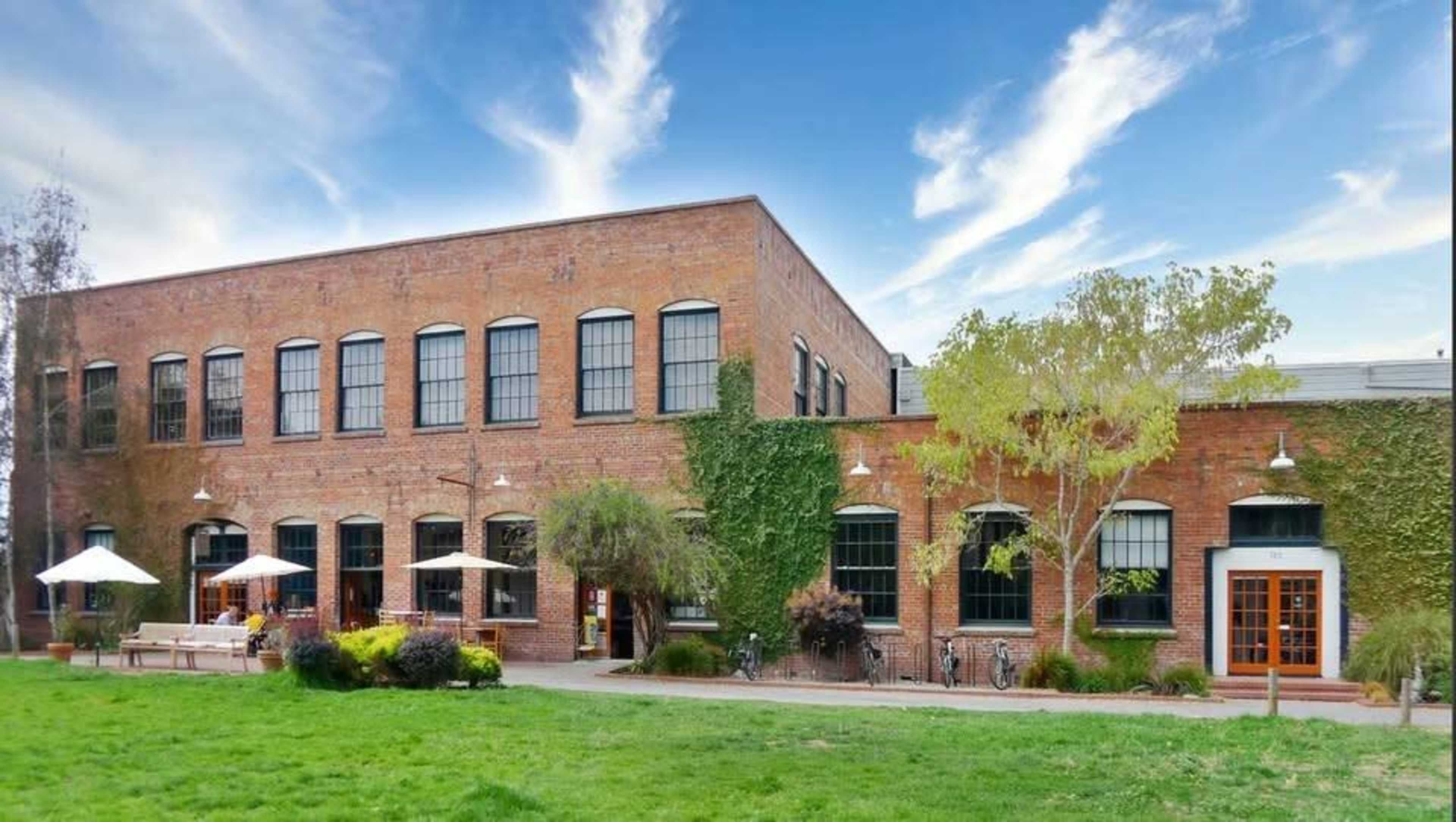 The image shows a red brick building surrounded by greenery, with outdoor seating areas and large windows.