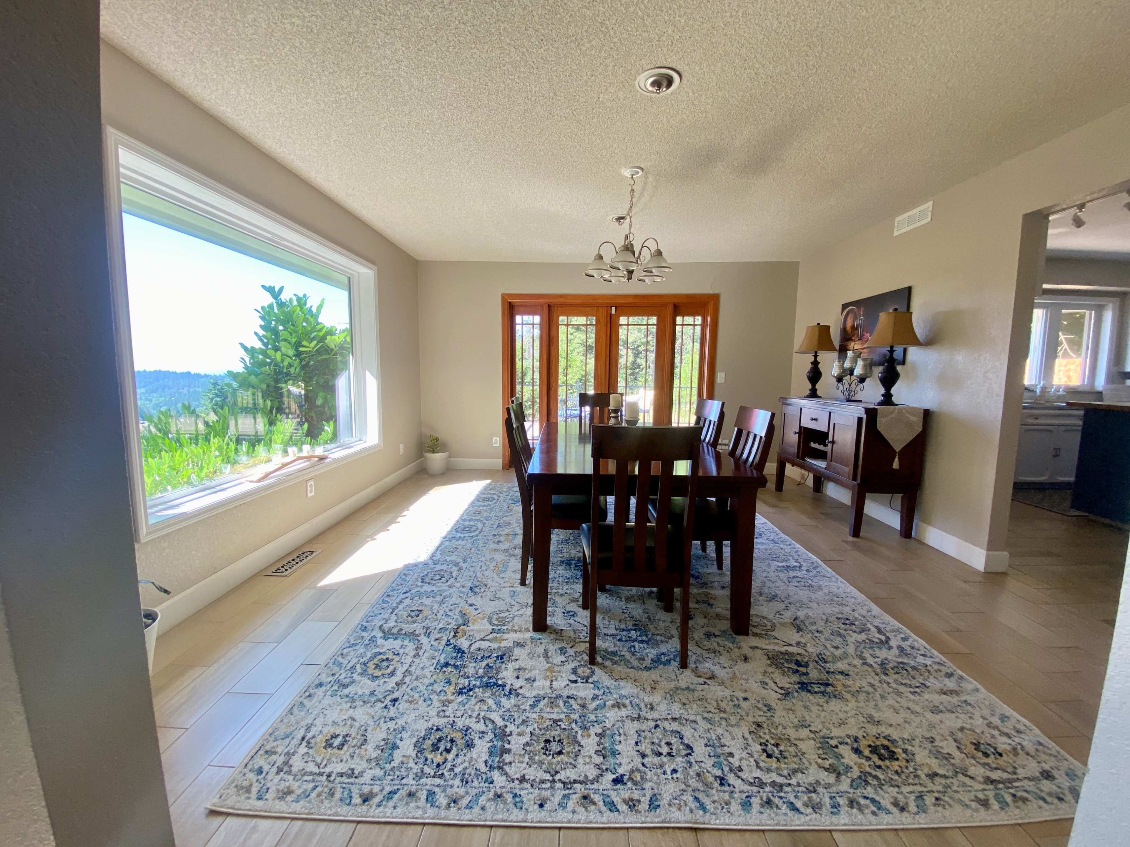 The image shows a dining room with a long wooden table, chairs, and a rug, featuring large windows that provide a view of a landscape outside.