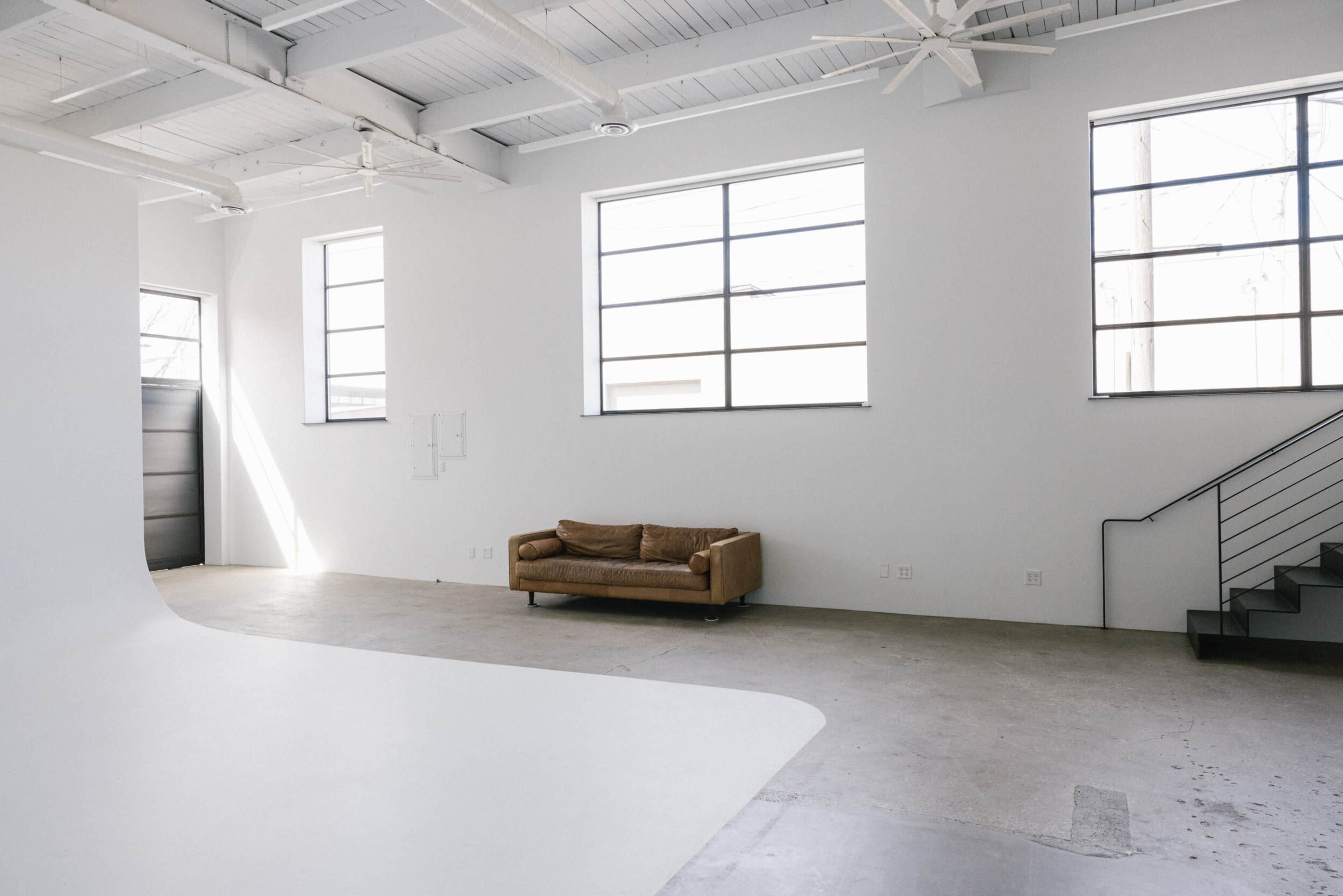 A spacious, minimalistic room with large windows, a brown couch, and a staircase along one wall.
