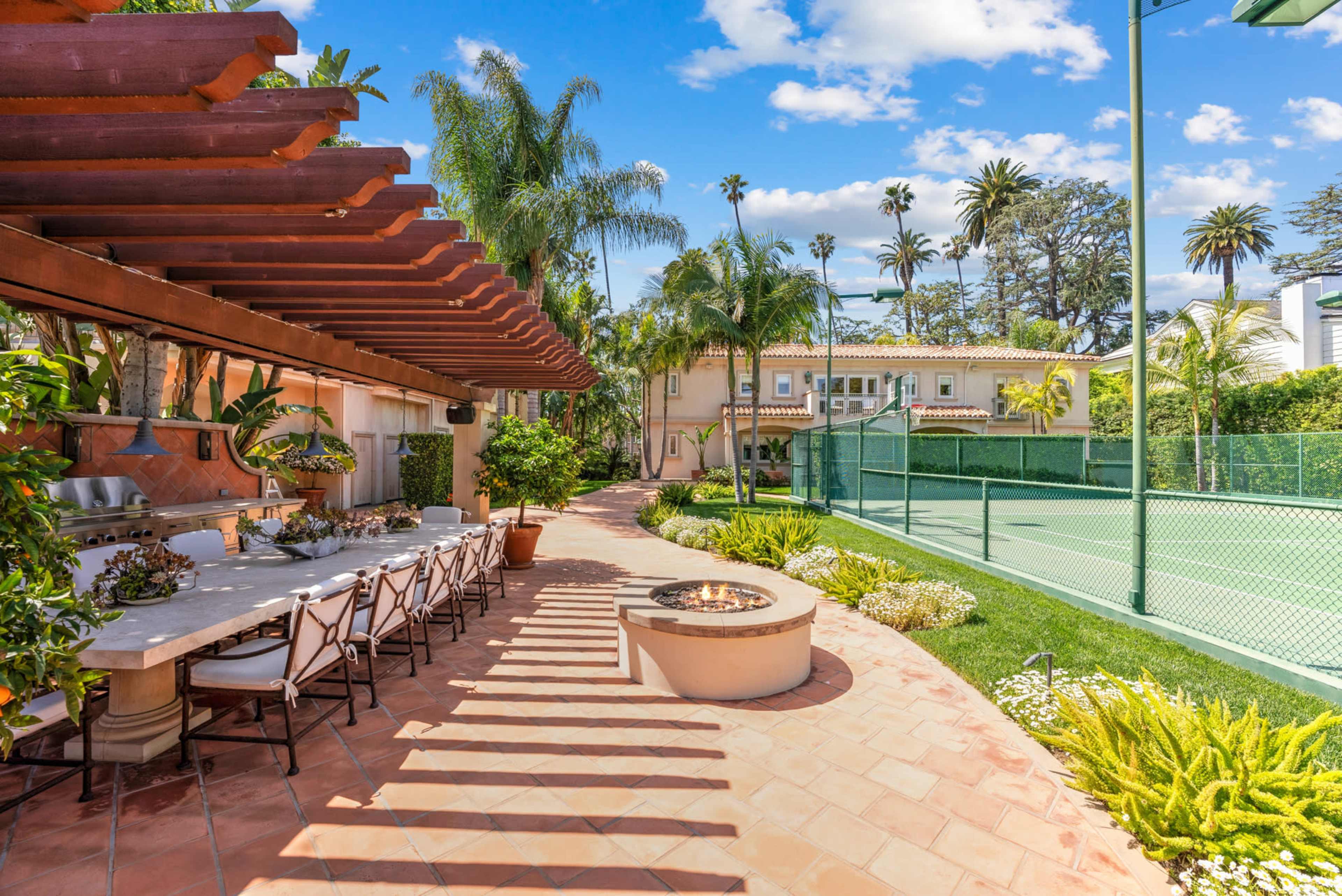 The image shows a landscaped backyard with a dining area under a pergola, surrounding greenery, and a fenced tennis court in the background.
