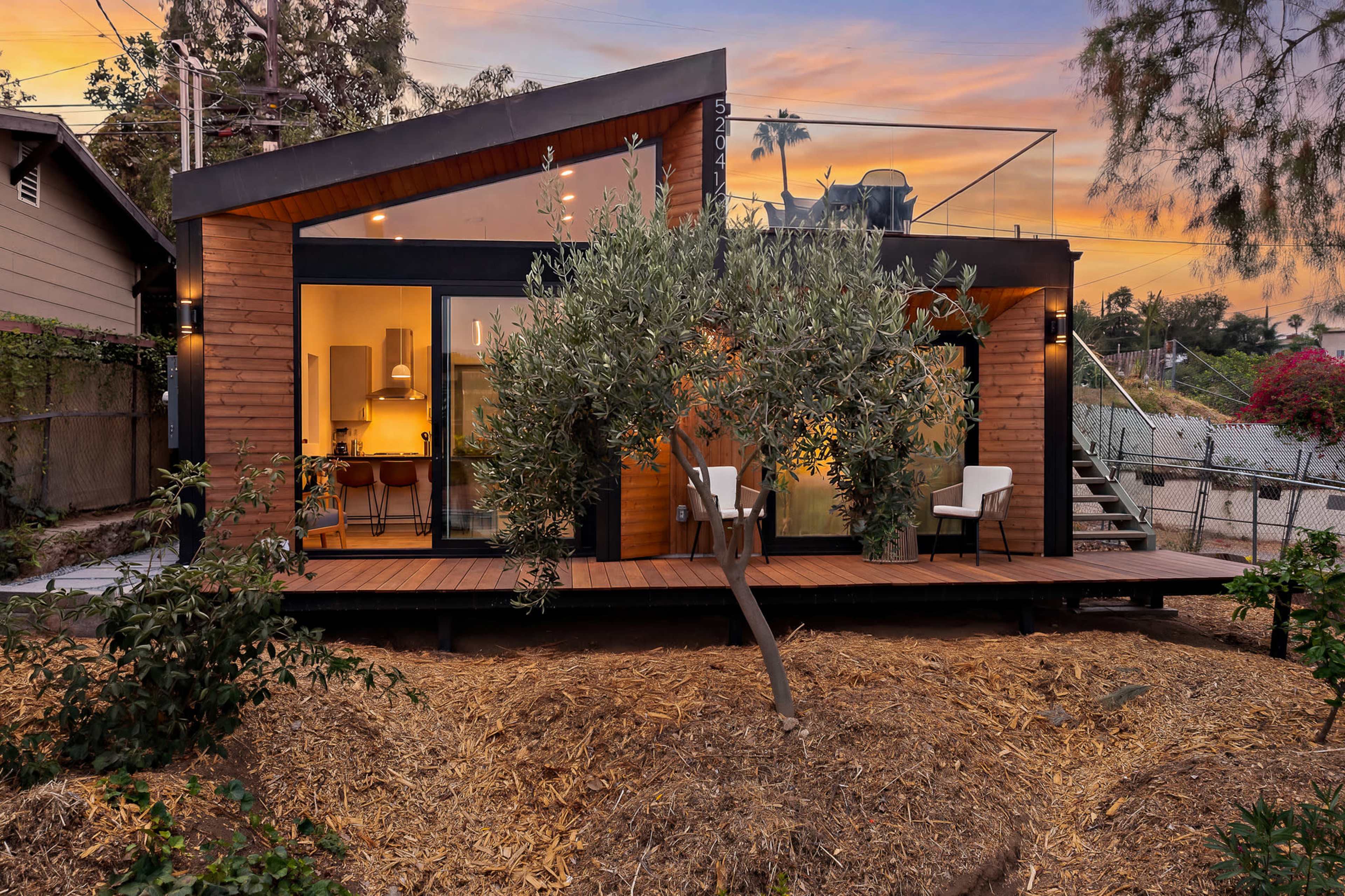 The image shows a modern wooden house with large glass windows, surrounded by greenery and a sloped landscape.