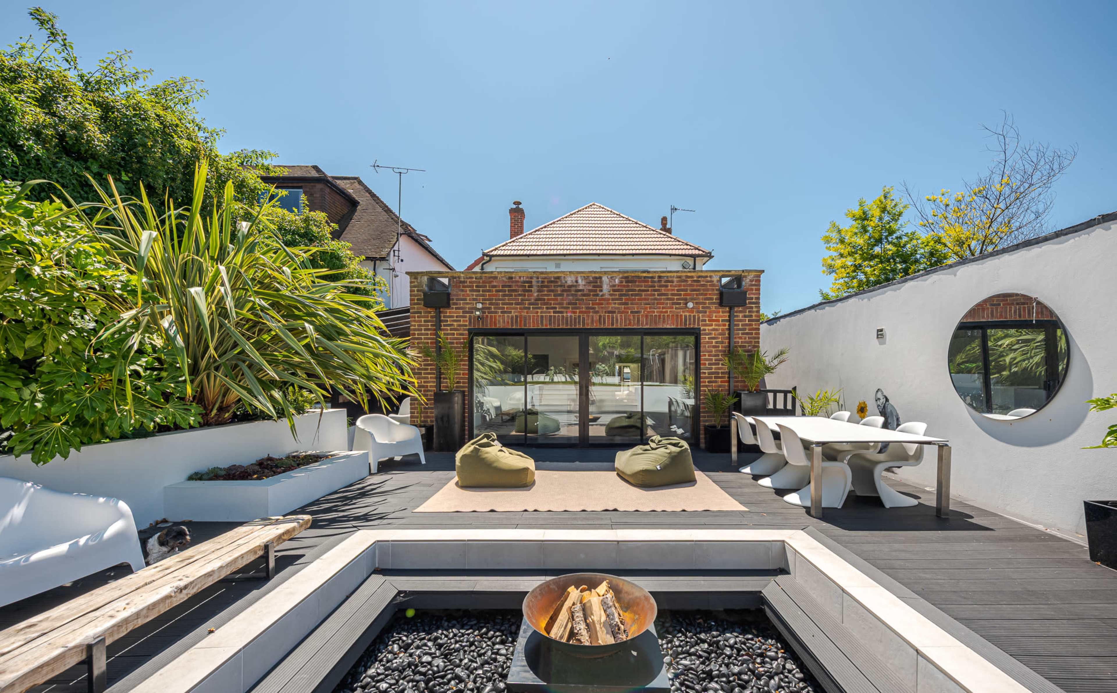 The image shows a modern outdoor patio area featuring seating, a fire pit, and lush greenery surrounding a glass-walled building.