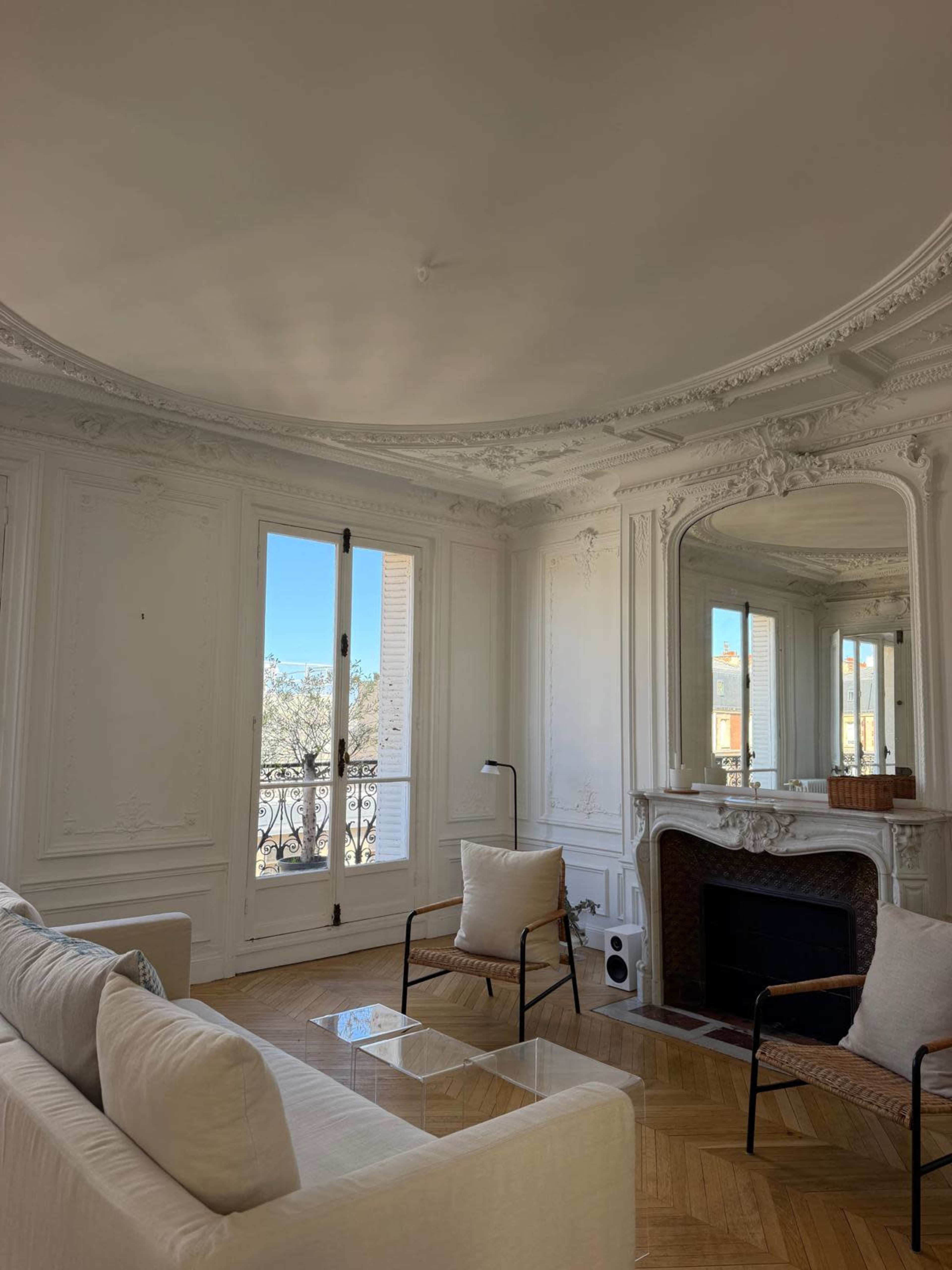 The image shows a elegantly designed living room featuring a large window with a view, a sofa, and two armchairs, with ornate architectural details on the walls and ceiling.