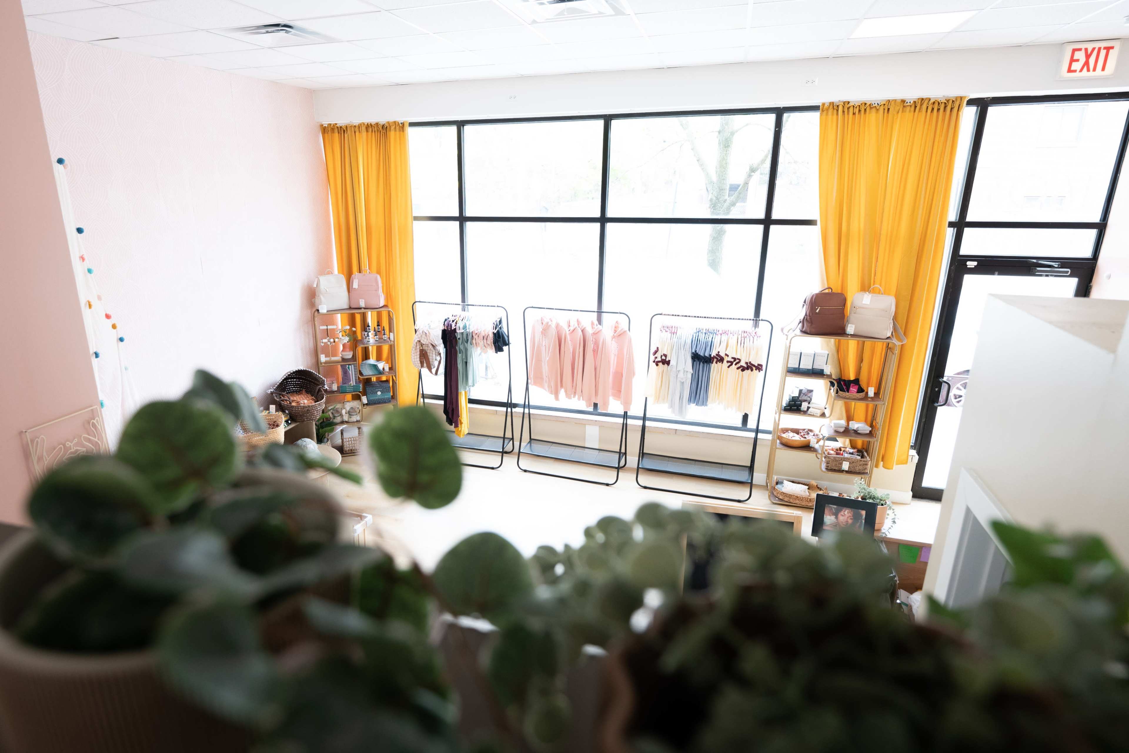 A boutique with clothing racks displaying various garments, large windows with yellow curtains, and shelves filled with accessories and plants.