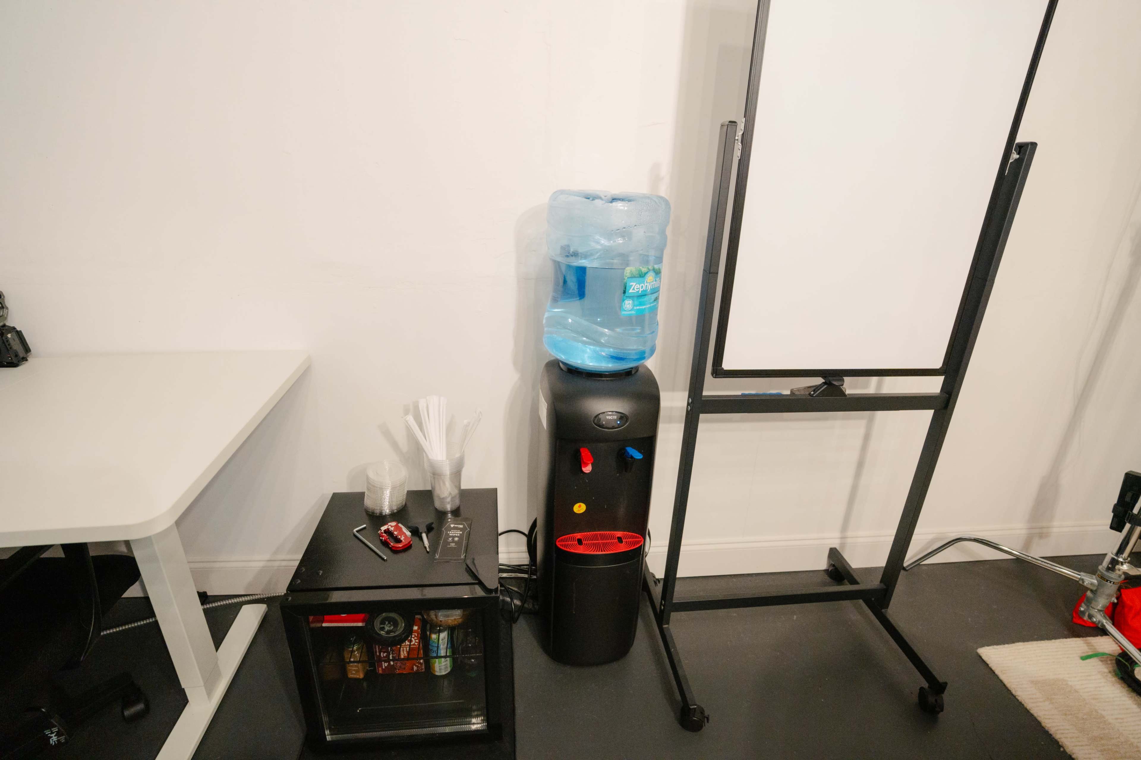 The image shows a water cooler with a blue jug, a small black fridge, and a whiteboard stand in a room.