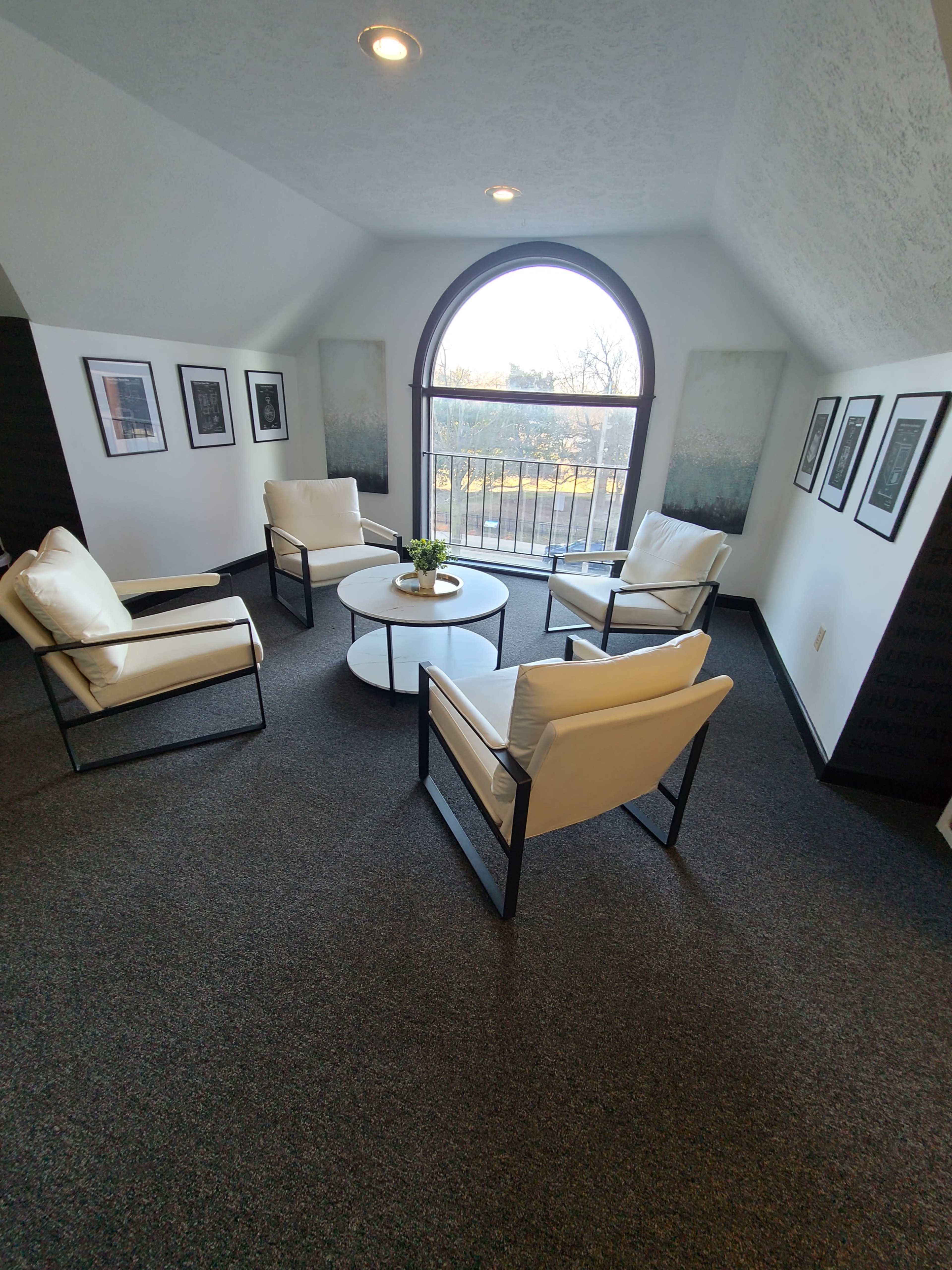 A modern sitting area features four white chairs surrounding a circular table, with framed photographs on the walls and a large arched window allowing natural light.