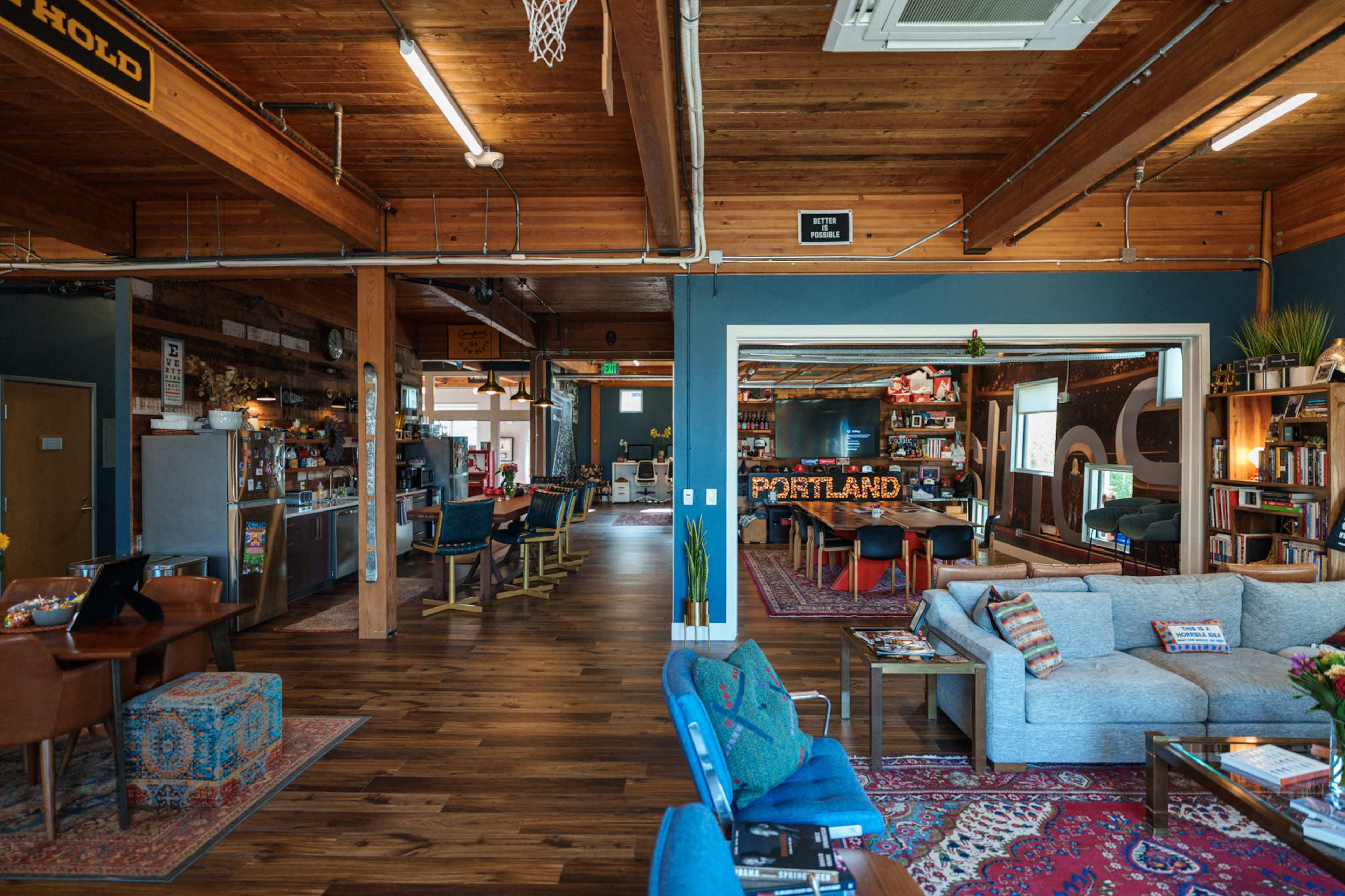 The image depicts a spacious, modern communal area with wooden beams, cozy seating arrangements, and shelves filled with books and decorations.