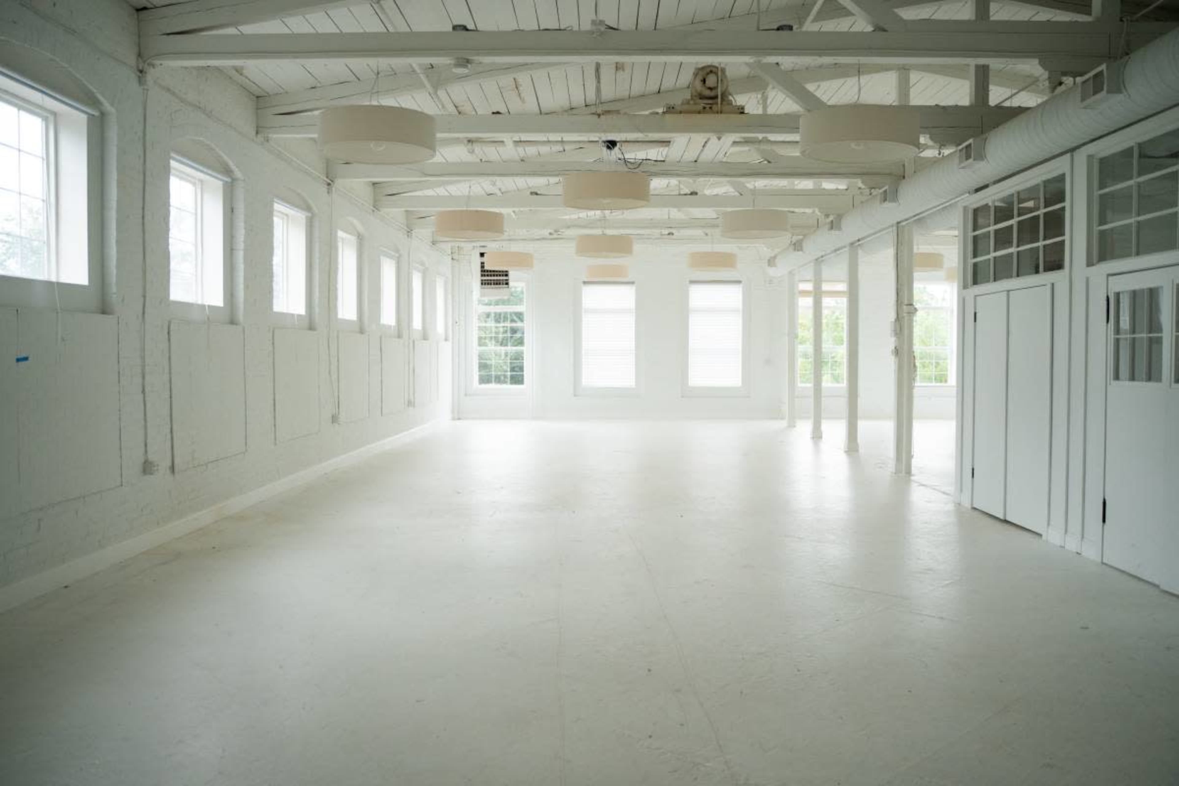 The image shows a spacious, empty room with white walls, large windows, and a minimalist design.