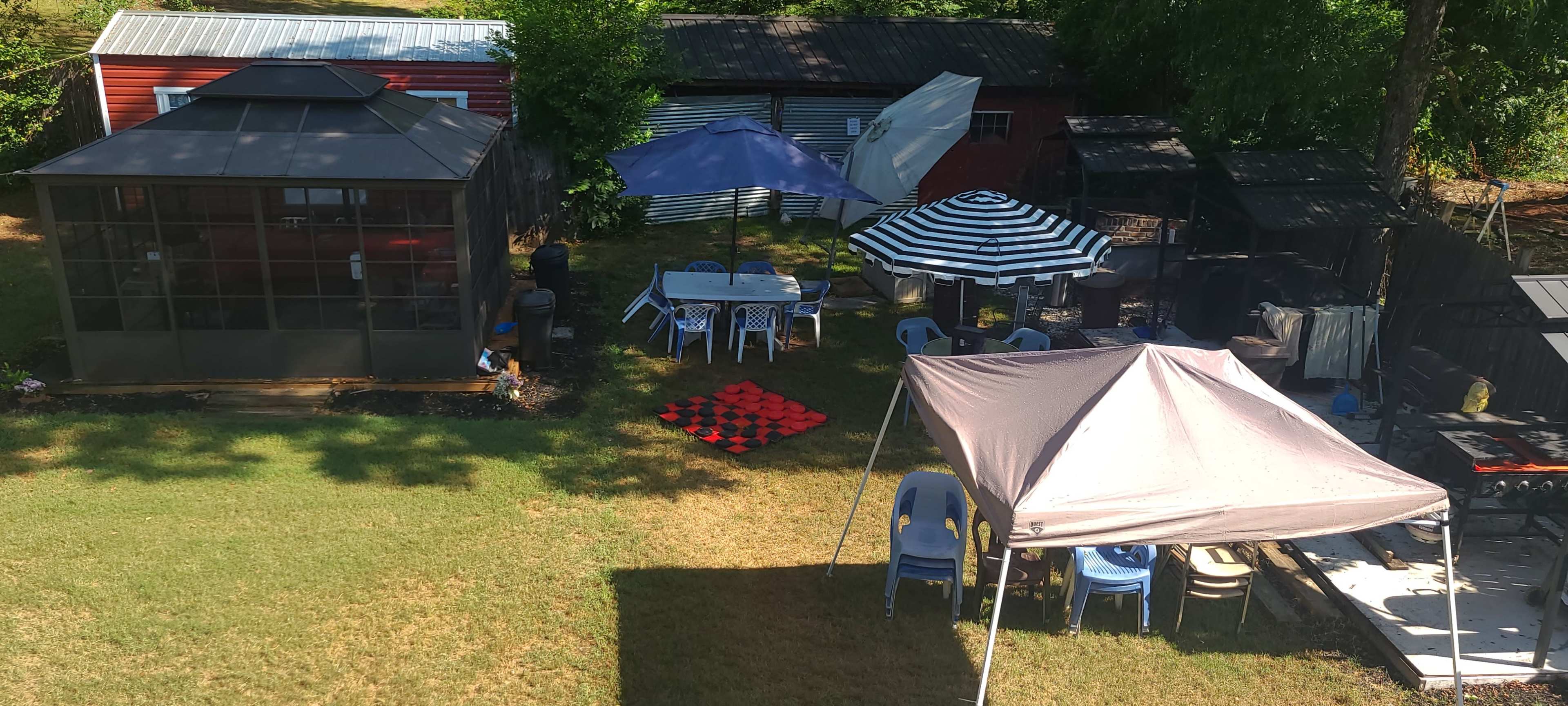 The image shows a backyard setup with various tents, seating areas, and game boards arranged on a lawn.