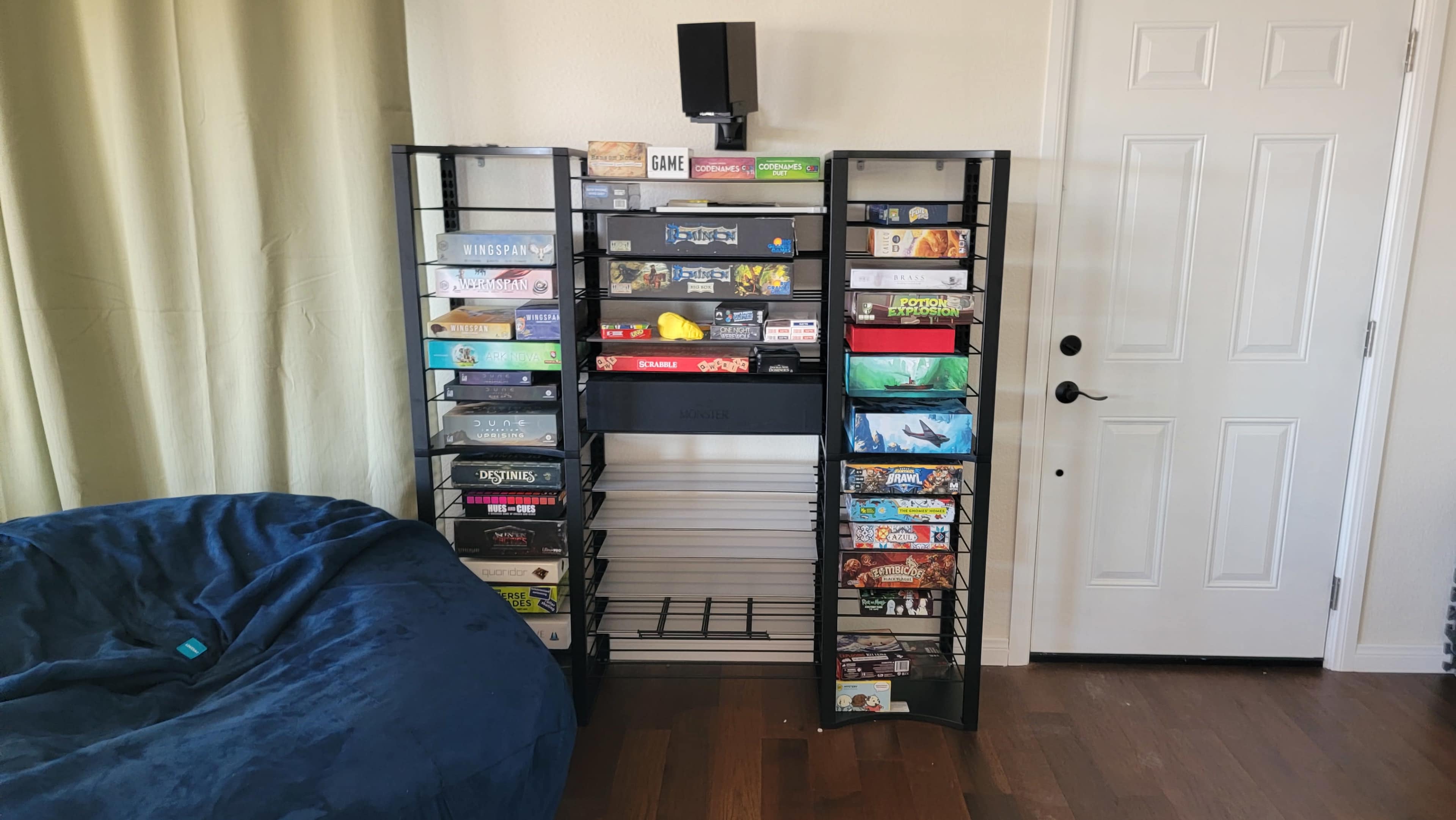 The image shows a black shelving unit filled with board games, alongside a blue bean bag chair and a door with a window.