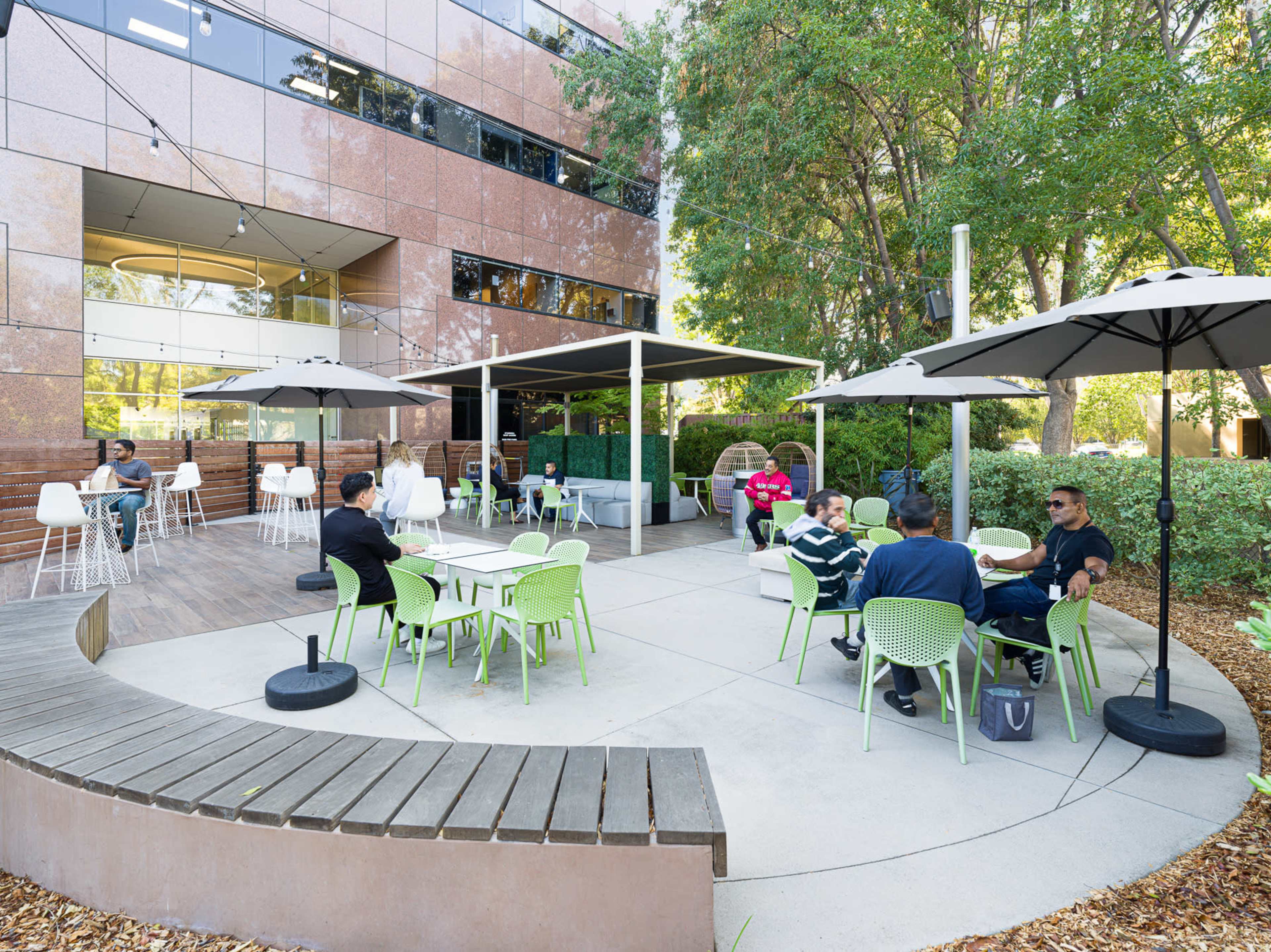 A group of people gathers at outdoor seating areas with green chairs and umbrellas near a modern building.