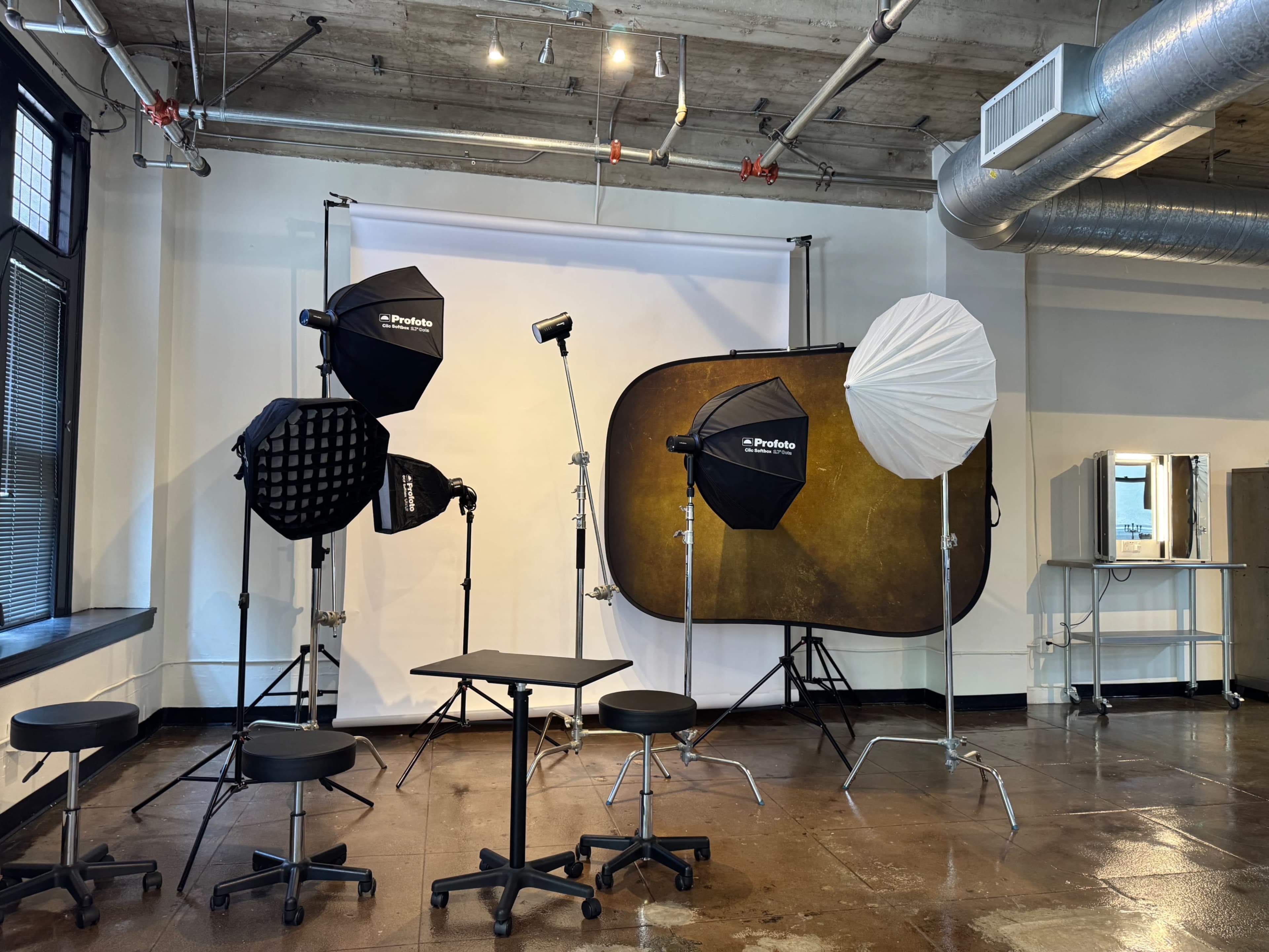 The image shows a photography studio set up with various lighting equipment, a white backdrop, and several chairs arranged around a small table.