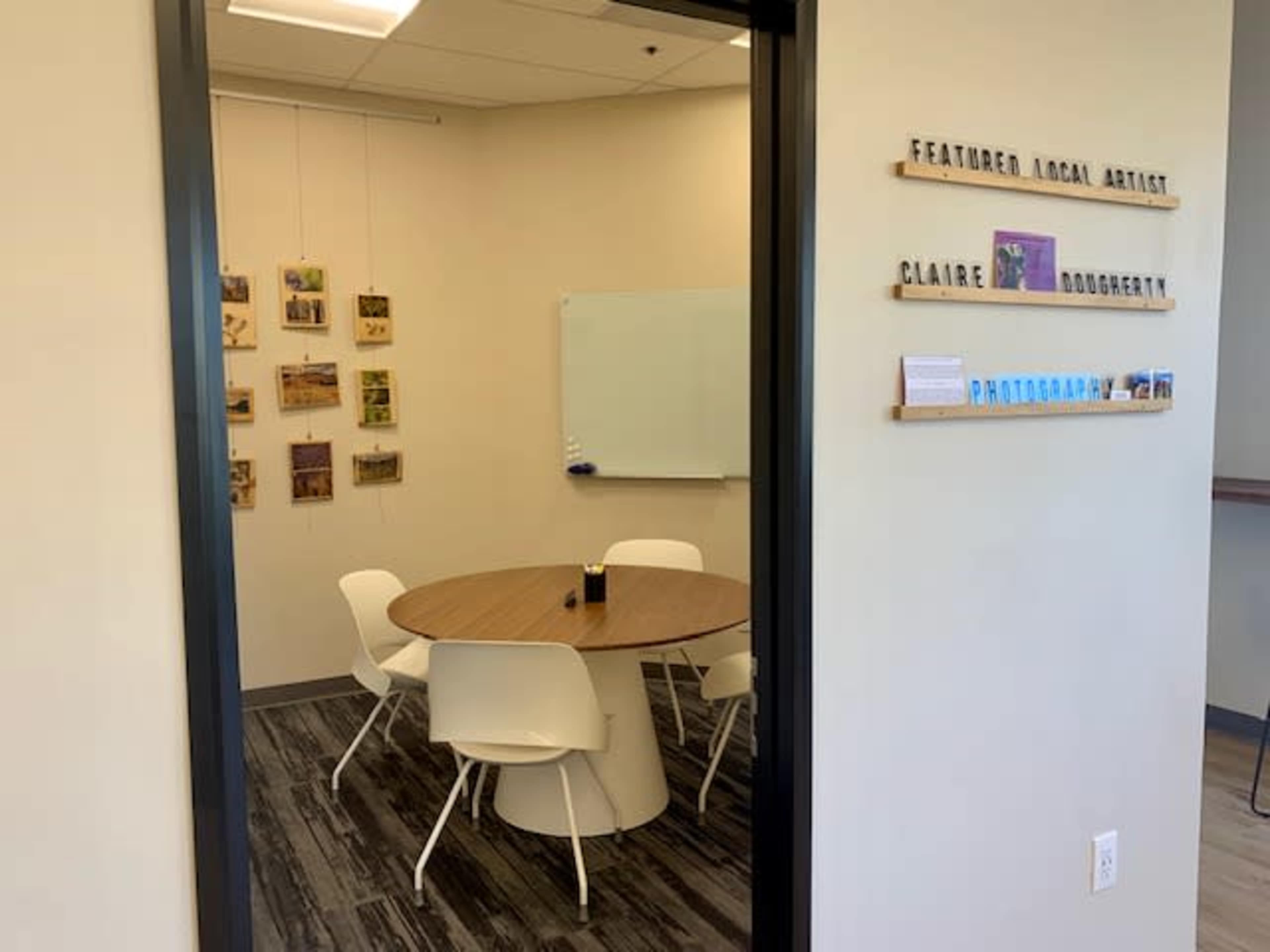 A small meeting room with a round wooden table surrounded by white chairs, featuring a gallery of photographs on the wall and a whiteboard.