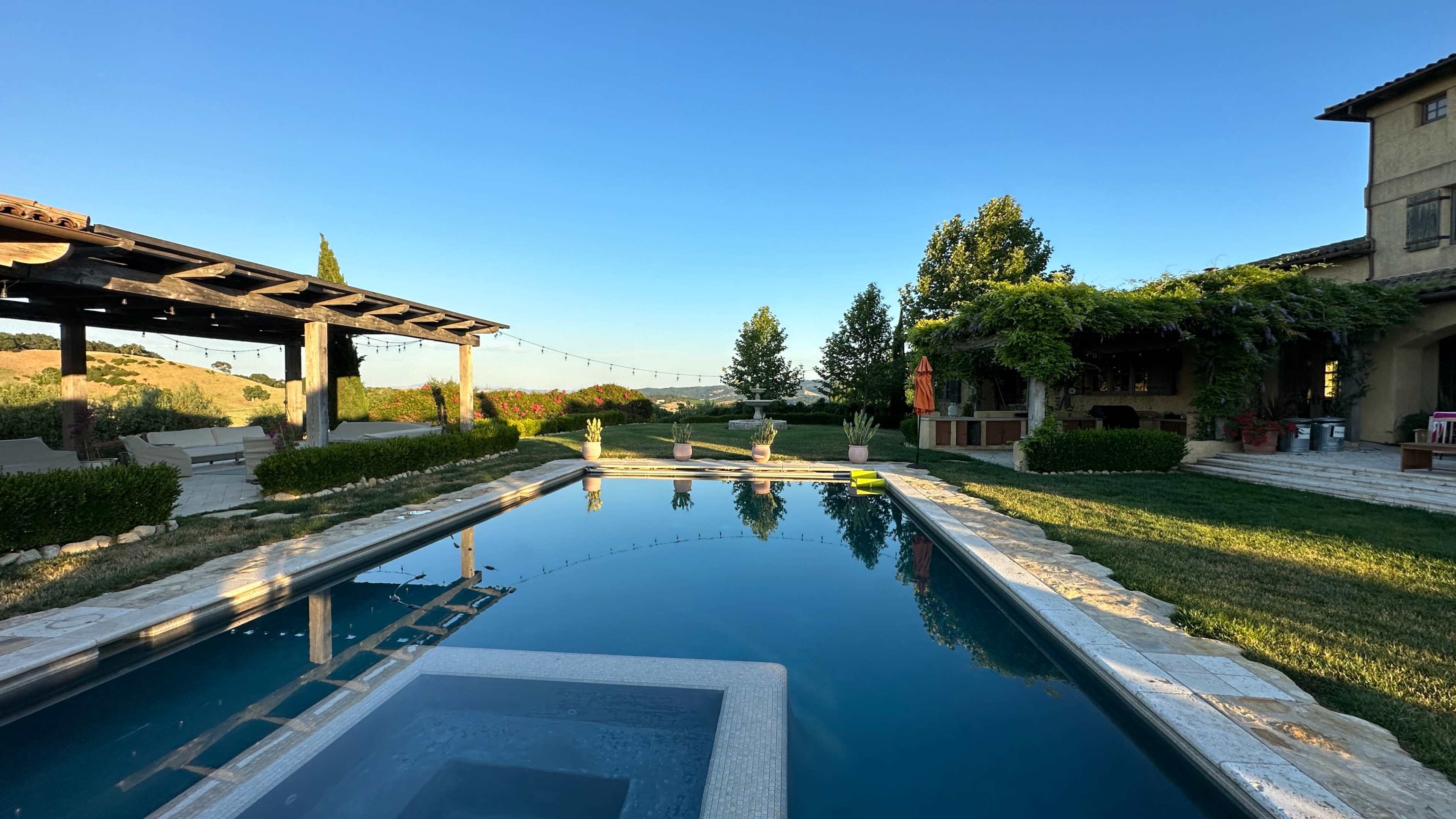 A clear swimming pool reflecting the blue sky, surrounded by landscaped gardens and a rustic patio area with wooden structures.